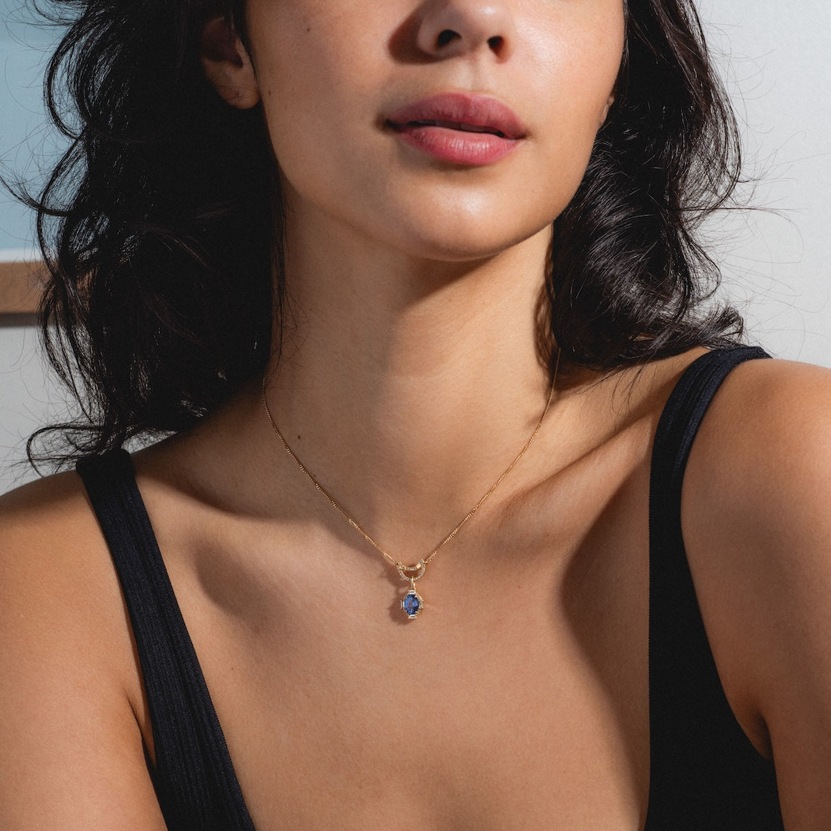 A woman wears the Solid Gold Perfect Start Charm Holder Necklace with a black sleeveless top, the delicate gold chain and blue gemstone pendant visible from the shoulders up in soft natural light.