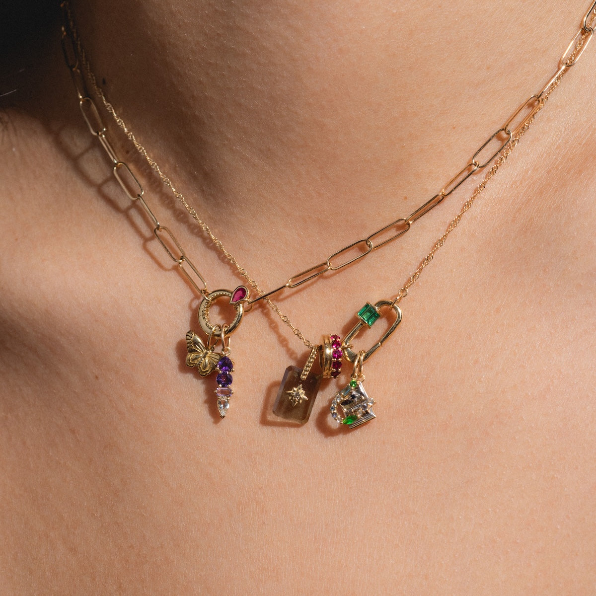Close-up of a person wearing the Solid Gold Perfect Start Charm Holder Necklace, layered stylishly with gold chains featuring unique pendants including gemstone charms, a green stone, and a lock-shaped pendant.