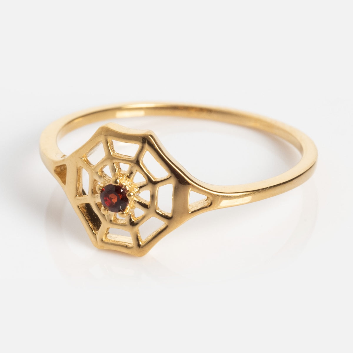 The Limited Edition Black Widow Garnet Spiderweb Ring features a gold spider web design with a small spider at the center, its body set with a red garnet gemstone, displayed on a white background.