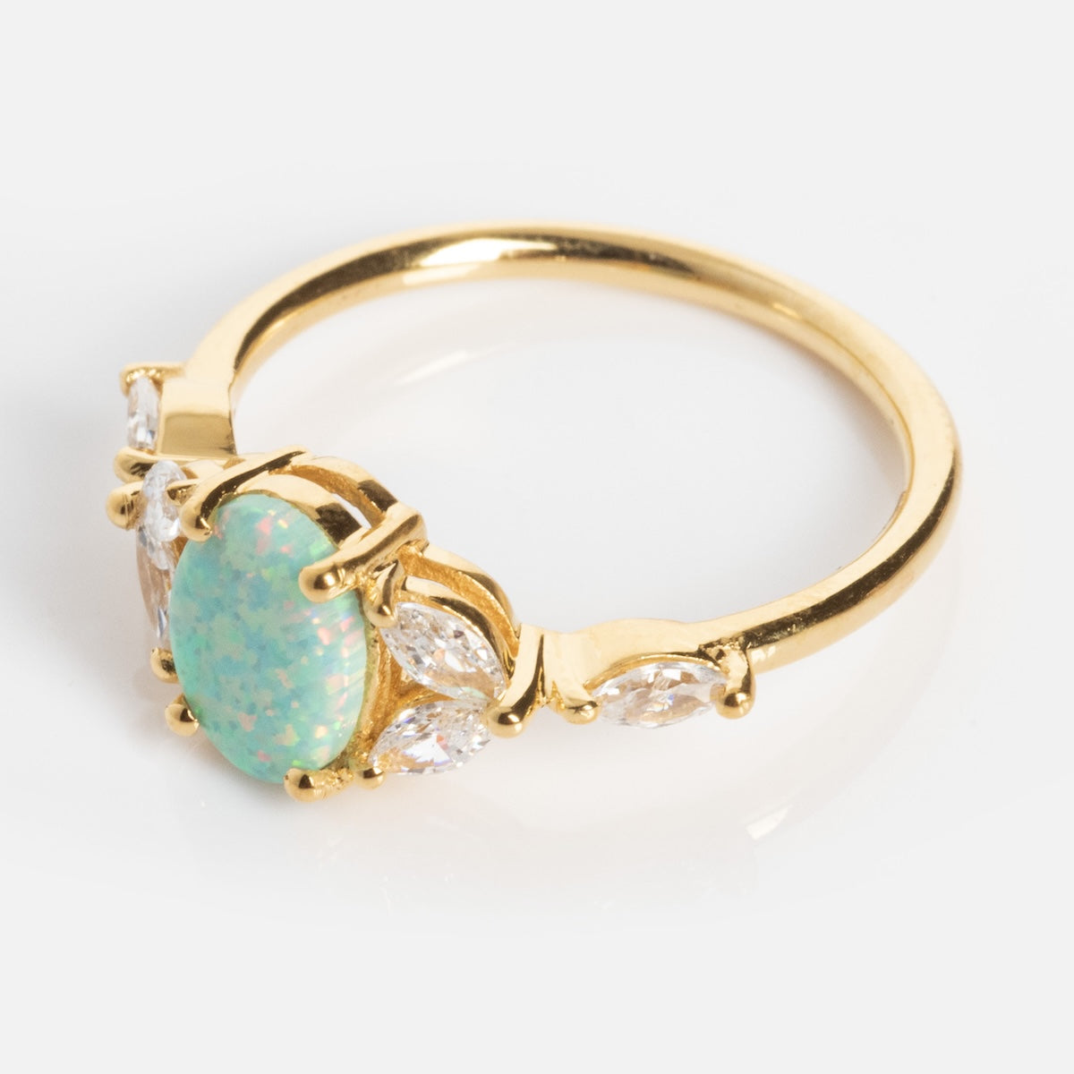 The Opal Enchanted Ring showcases an oval-shaped opal with green and blue hues, accented by marquise-cut clear stones on each side, all set in gold, displayed against a white background.
