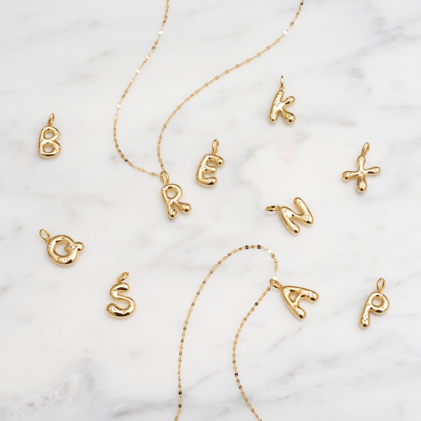Star Crossed Celestial Bubble Letter Charms in B, R, E, A, K, O, S, N, and P and two gold chains are displayed on a white marble surface.