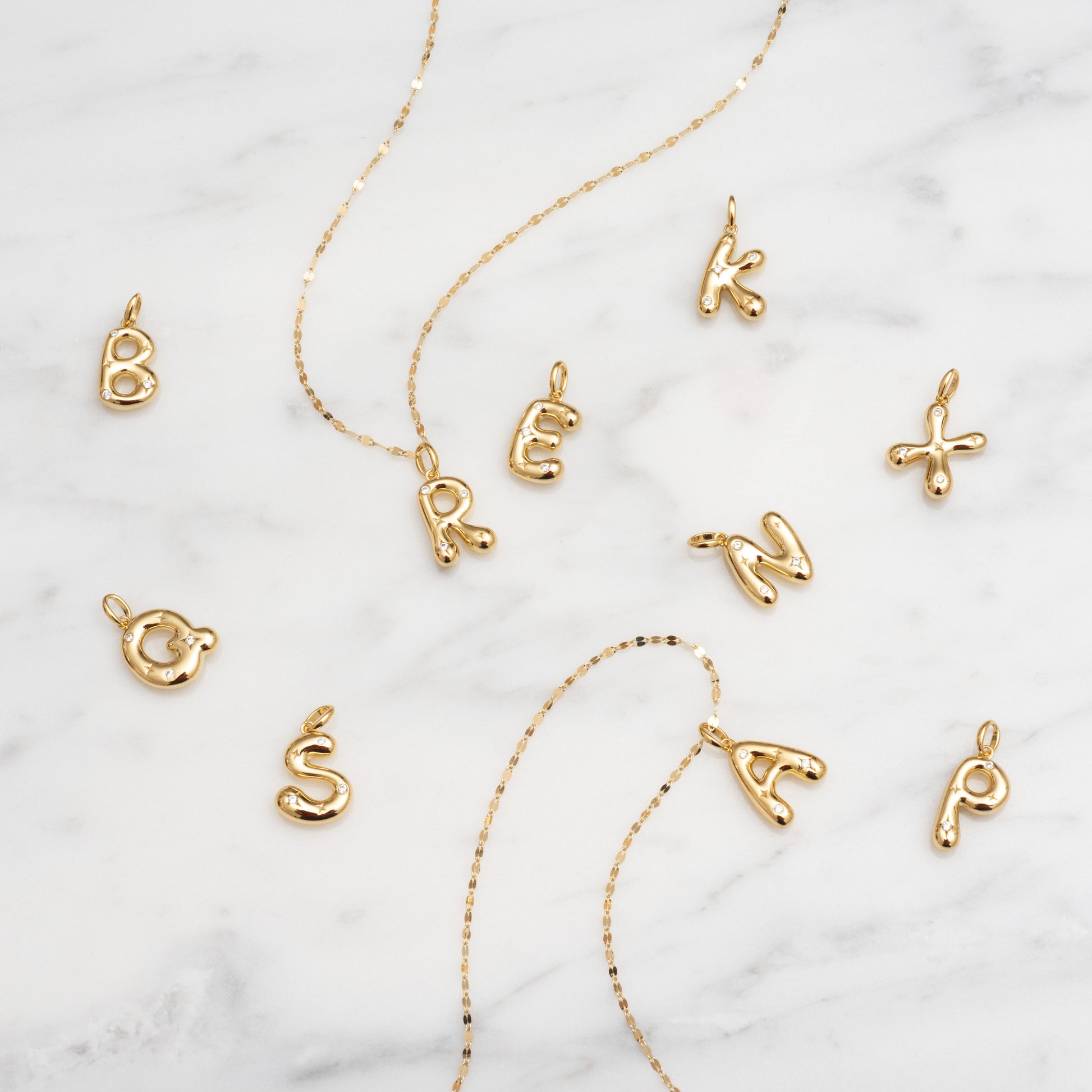 Star Crossed Celestial Bubble Letter Charms in B, R, E, A, K, O, S, N, and P and two gold chains are displayed on a white marble surface.