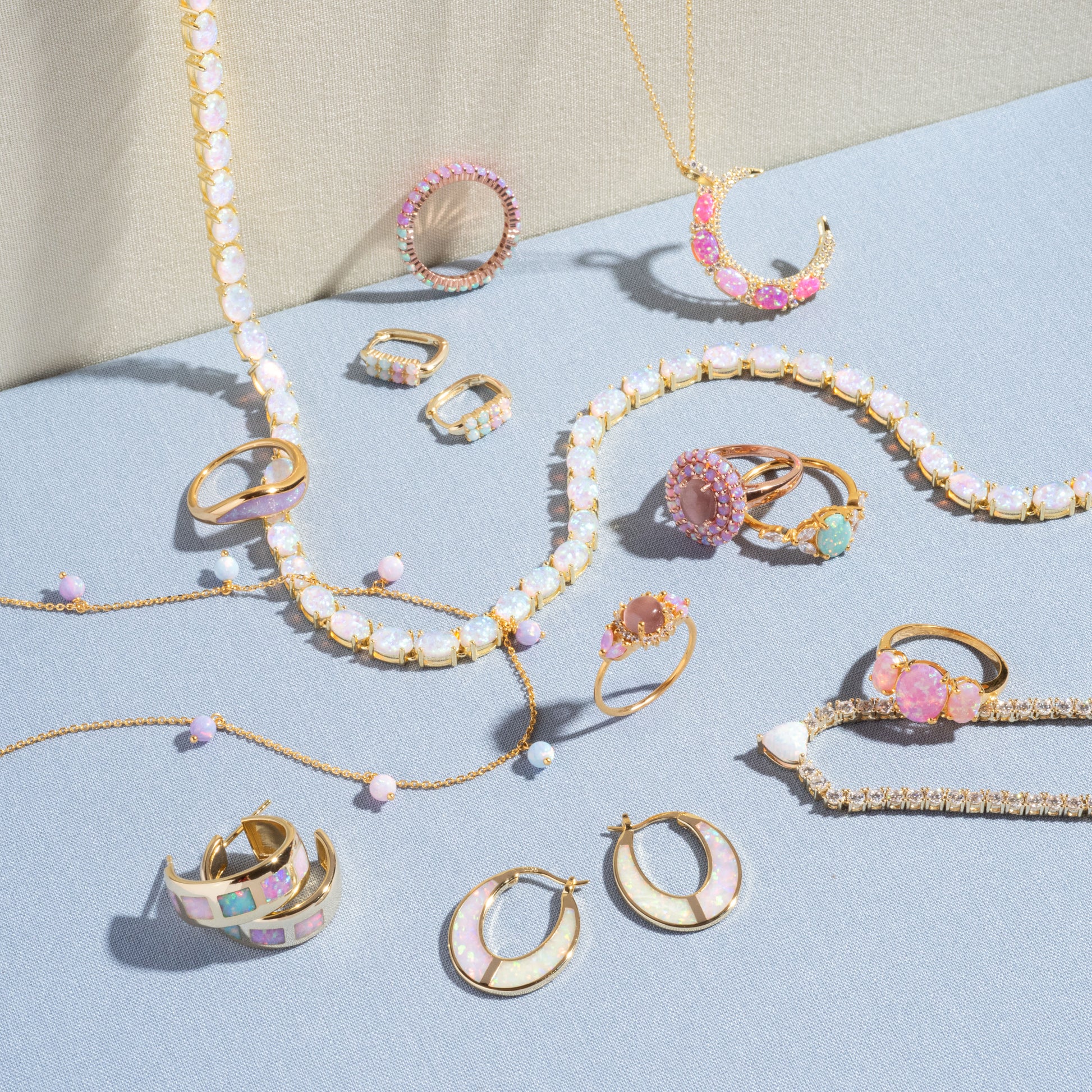 The Opal Dreamers Wave Ring, a gold ring with pink, white, and pastel gemstones, is displayed on light blue and beige fabric backgrounds along with other jewelry pieces.