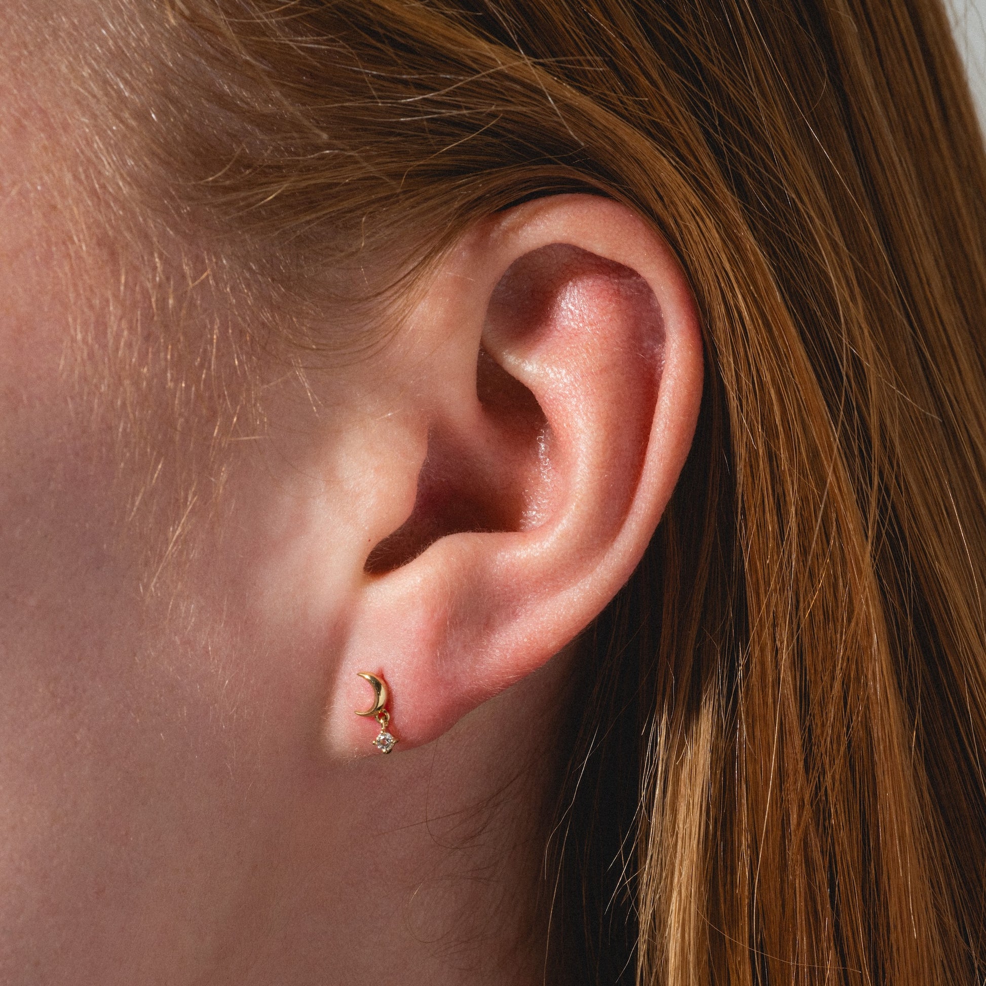 Close-up of a person's ear with straight, reddish-brown hair wearing Solid Gold Crescent Moon Flat Back Studs featuring a small clear gemstone. The softly lit background is out of focus.