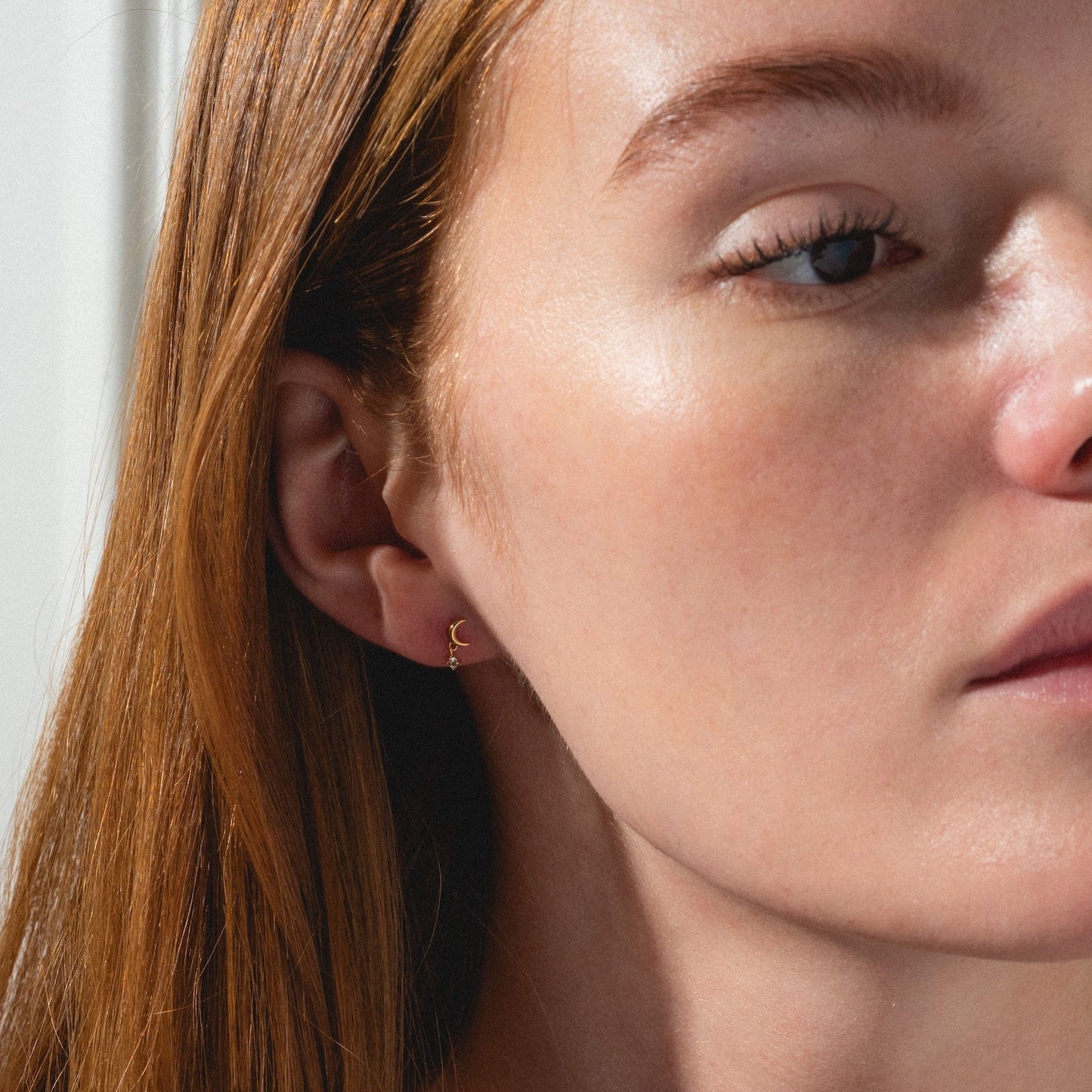 A close-up of a woman with long red hair and fair skin, wearing Solid Gold Crescent Moon Flat Back Studs. She has natural makeup with defined brows, and the lighting highlights her smooth skin as she looks slightly to the side.