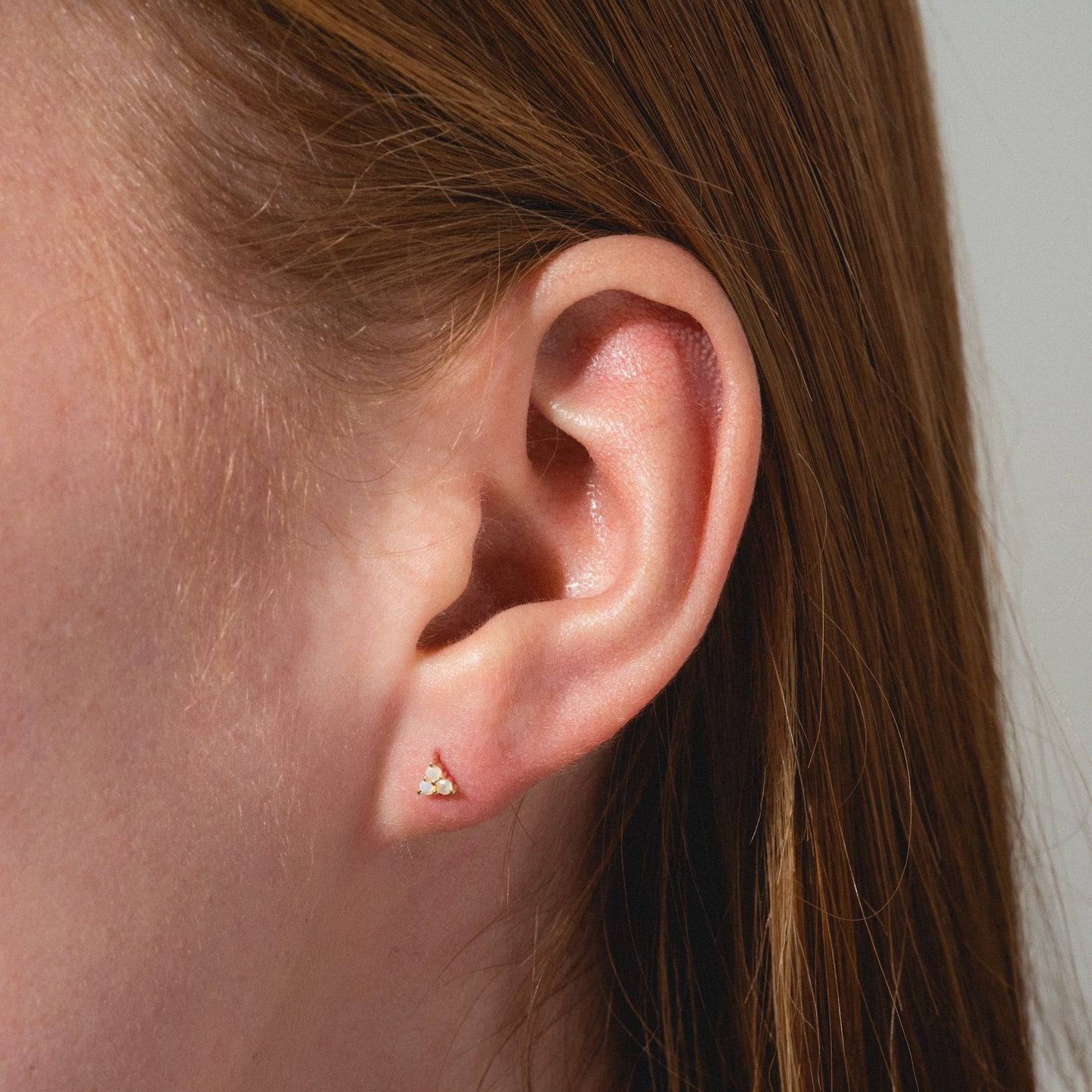Close-up of a person's ear with straight, light brown hair wearing the Solid Gold Opal Trio Flat Back Studs—delicate earrings featuring three opals in a triangular pattern. The neutral background is softly blurred.