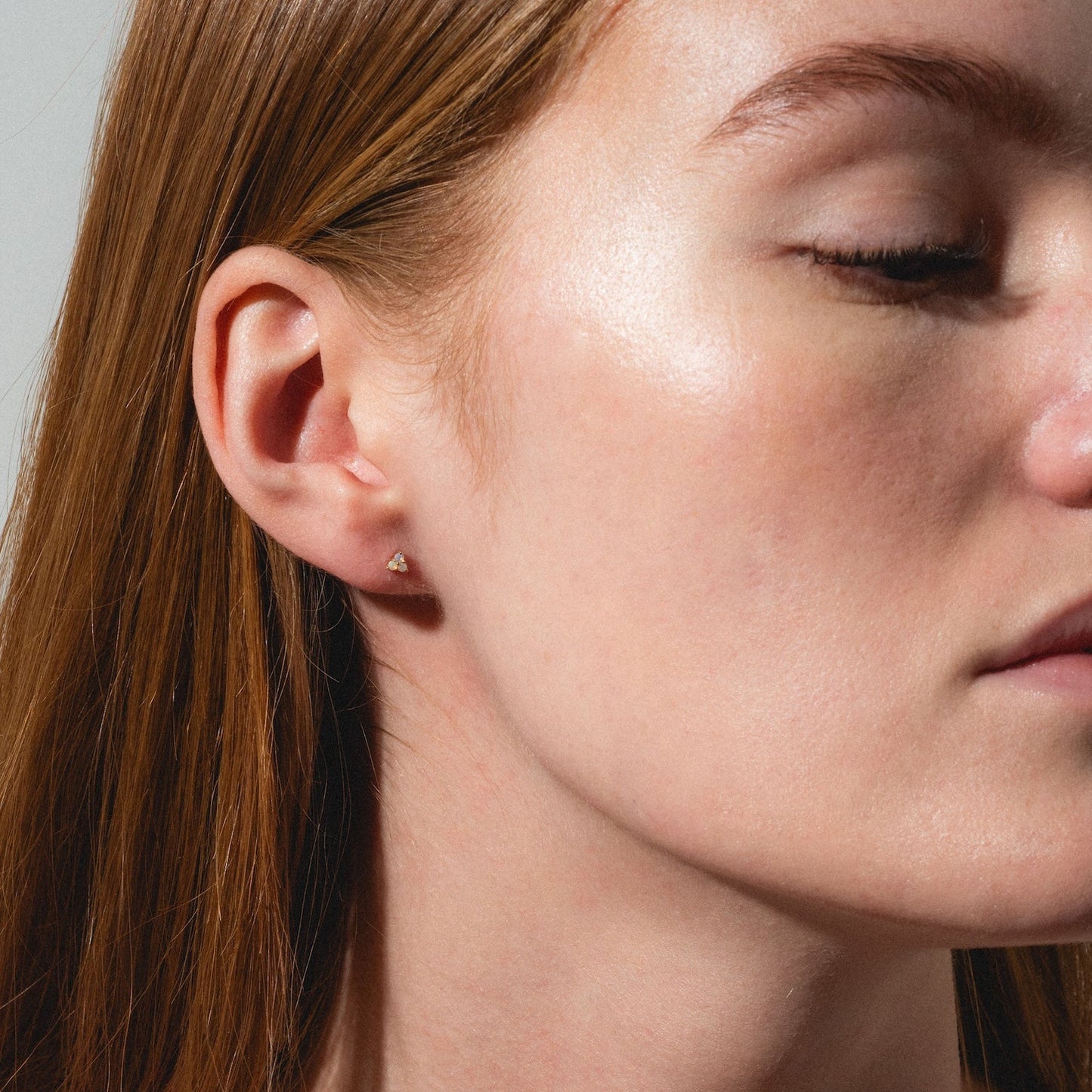 A close-up of a woman with long red hair and fair skin, wearing Solid Gold Opal Trio Flat Back Studs, eyes closed and head slightly turned to the side against a neutral background.
