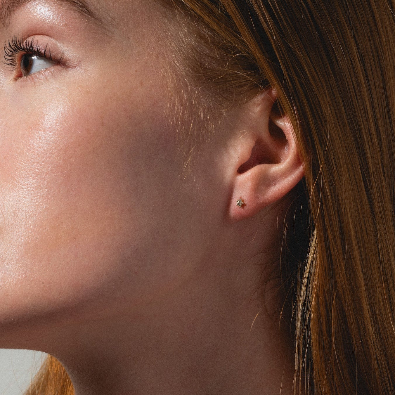 A woman with long red hair and fair skin looks to the left, wearing Solid Gold Sparkling Star Flat Back Studs and a black thin-strapped top against a plain, light background.