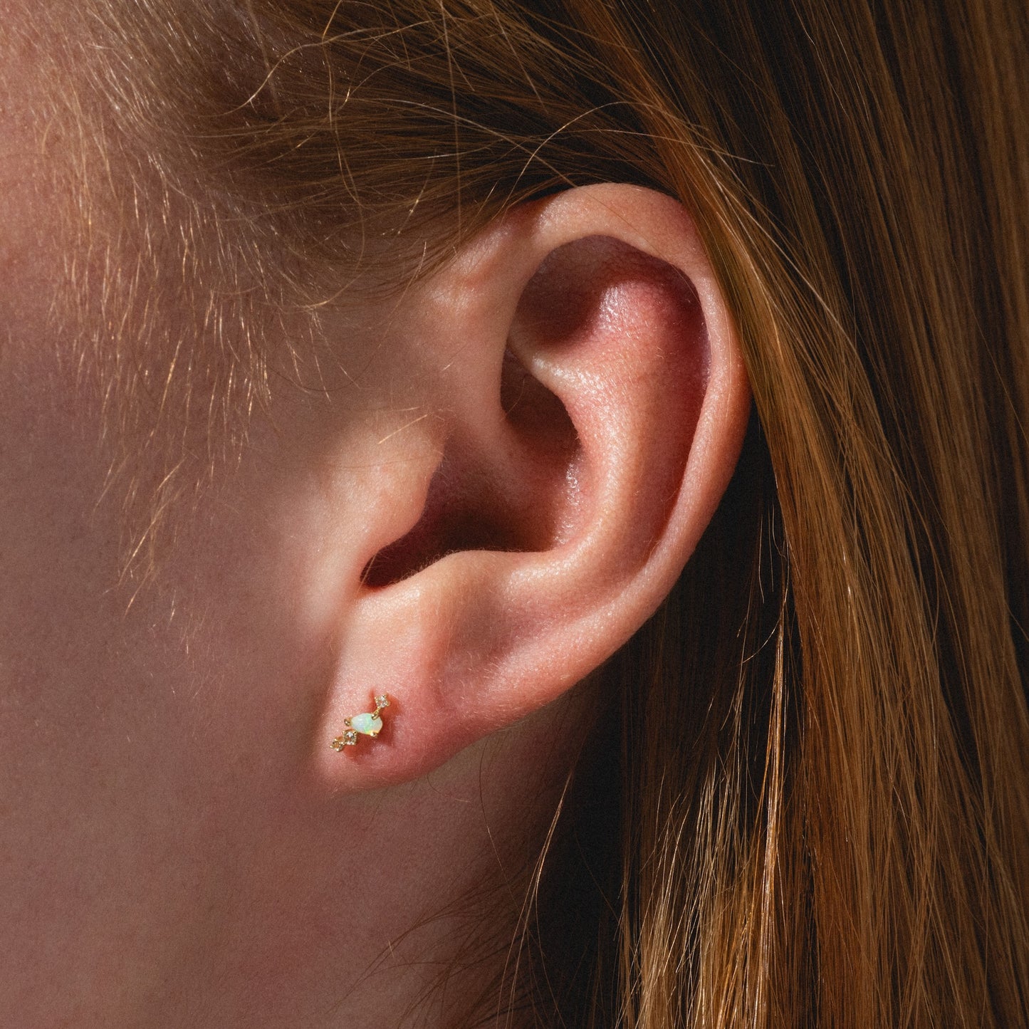 A close-up of a person’s ear with straight red hair, wearing Solid Gold Opal Constellation Flat Back Studs featuring a light green gemstone.