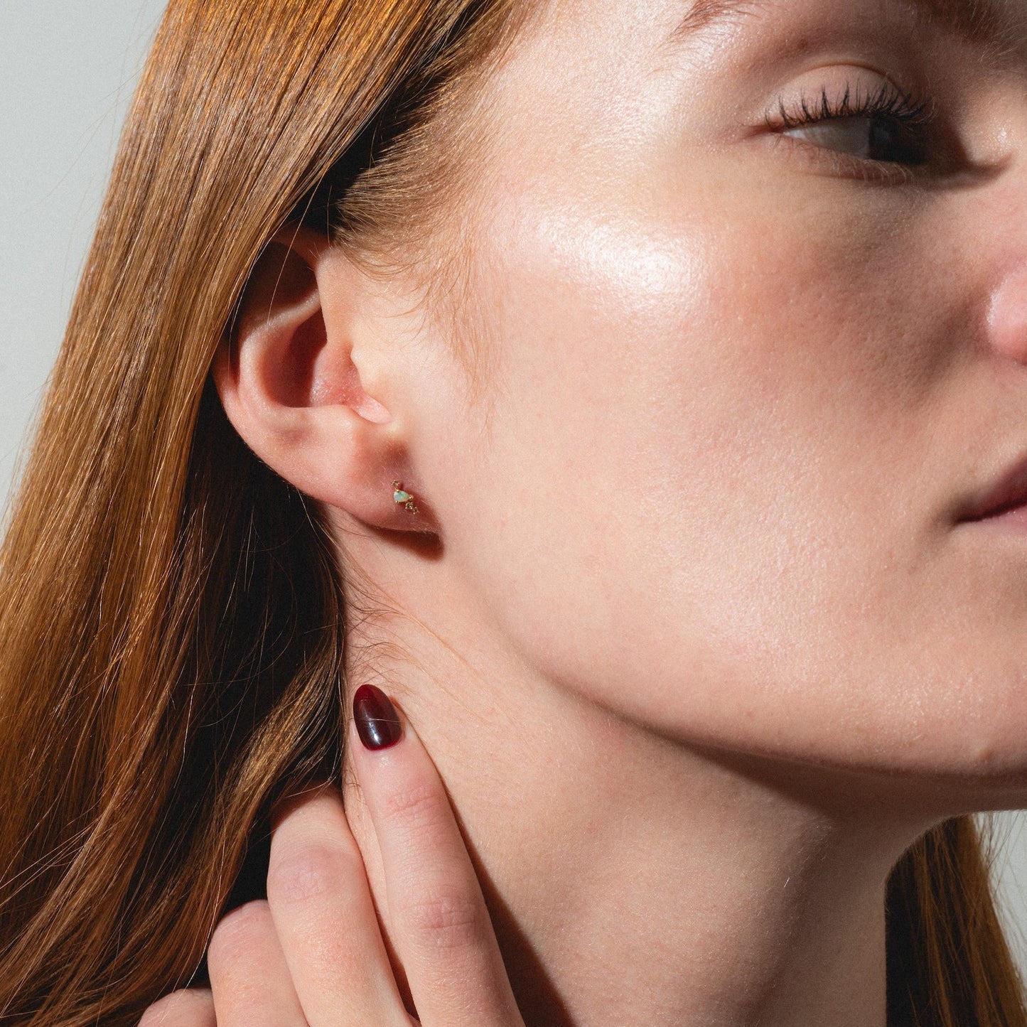 A close-up of a woman with fair skin and straight red hair gently touching her neck, showcasing Solid Gold Opal Constellation Flat Back Studs. She has glossy, clear skin and natural makeup.