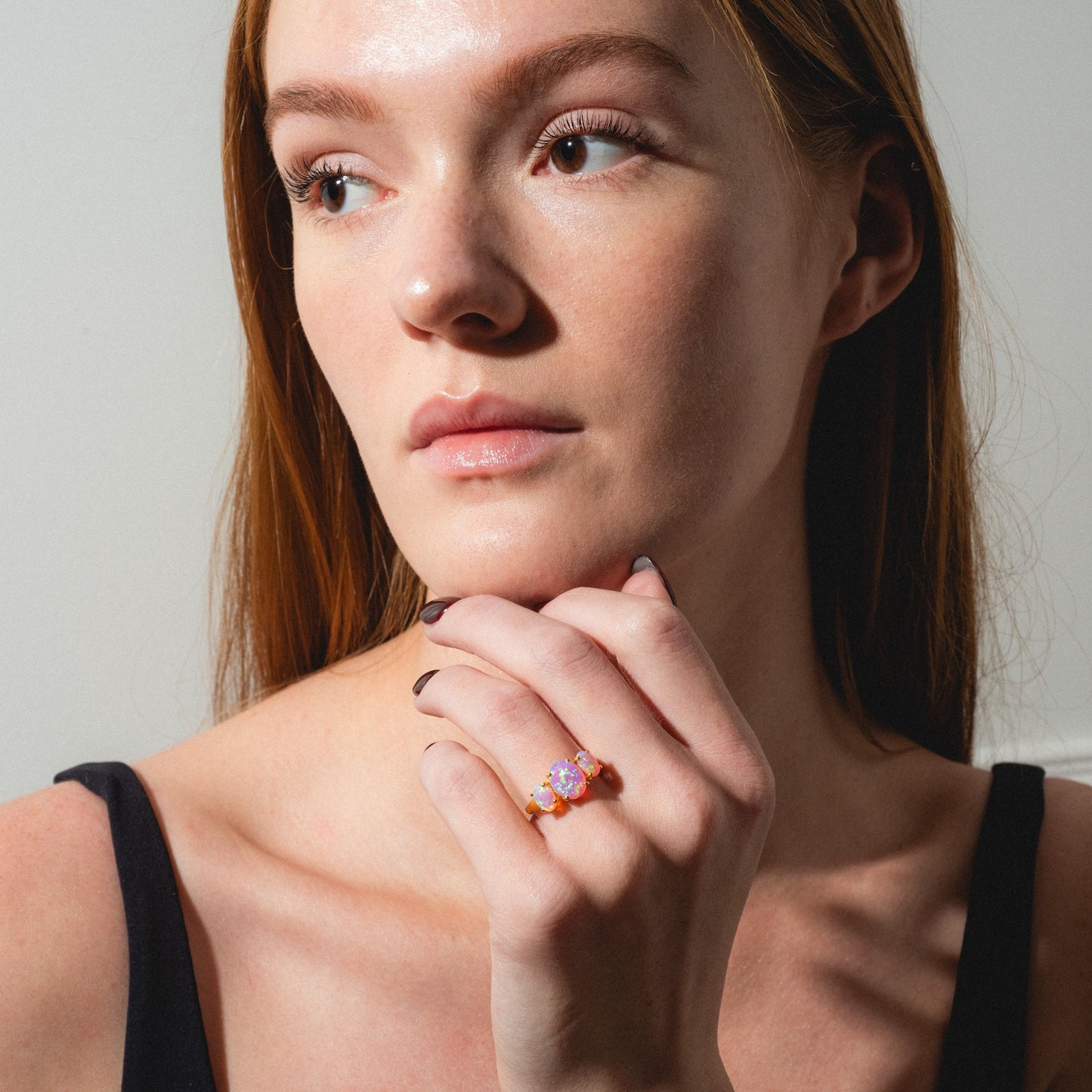 A fair-skinned woman with long red hair gazes thoughtfully to the side, her chin resting on her hand to display the Pink Opal Gum Drop Ring. She wears a black sleeveless top, illuminated by natural light.