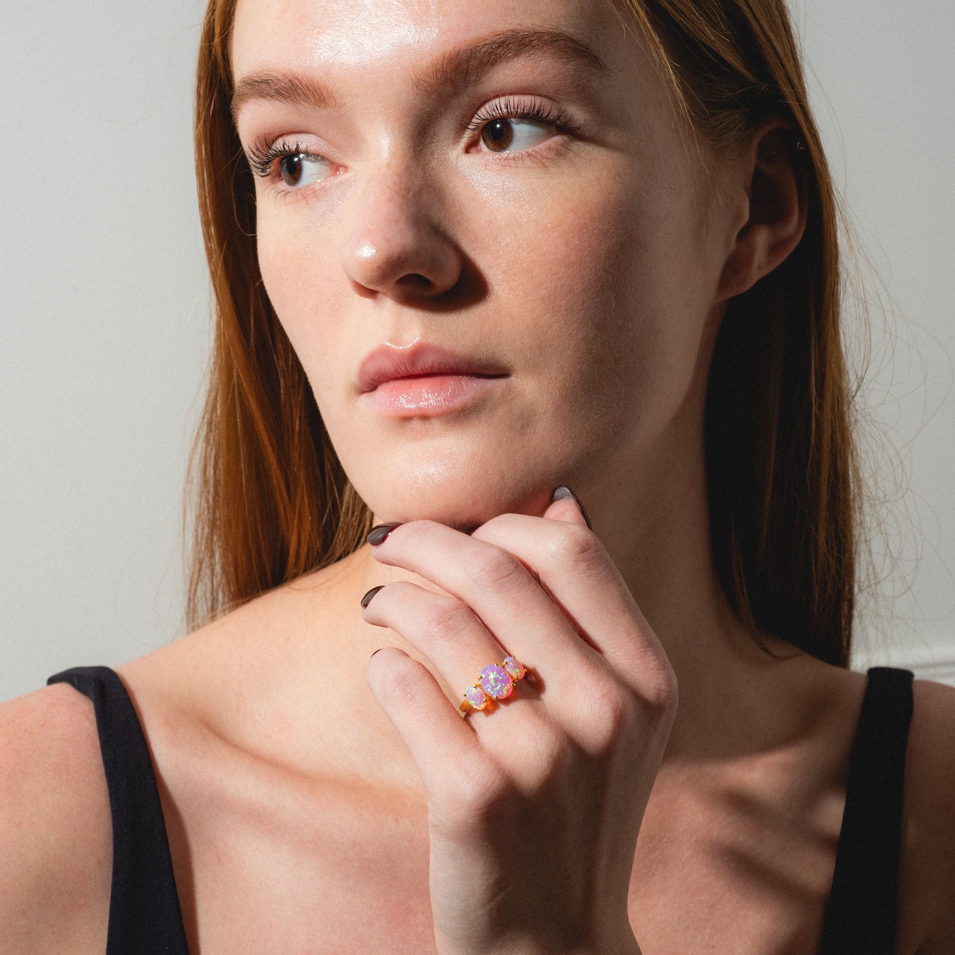 A fair-skinned woman with long red hair gazes thoughtfully to the side, her chin resting on her hand to display the Pink Opal Gum Drop Ring. She wears a black sleeveless top, illuminated by natural light.