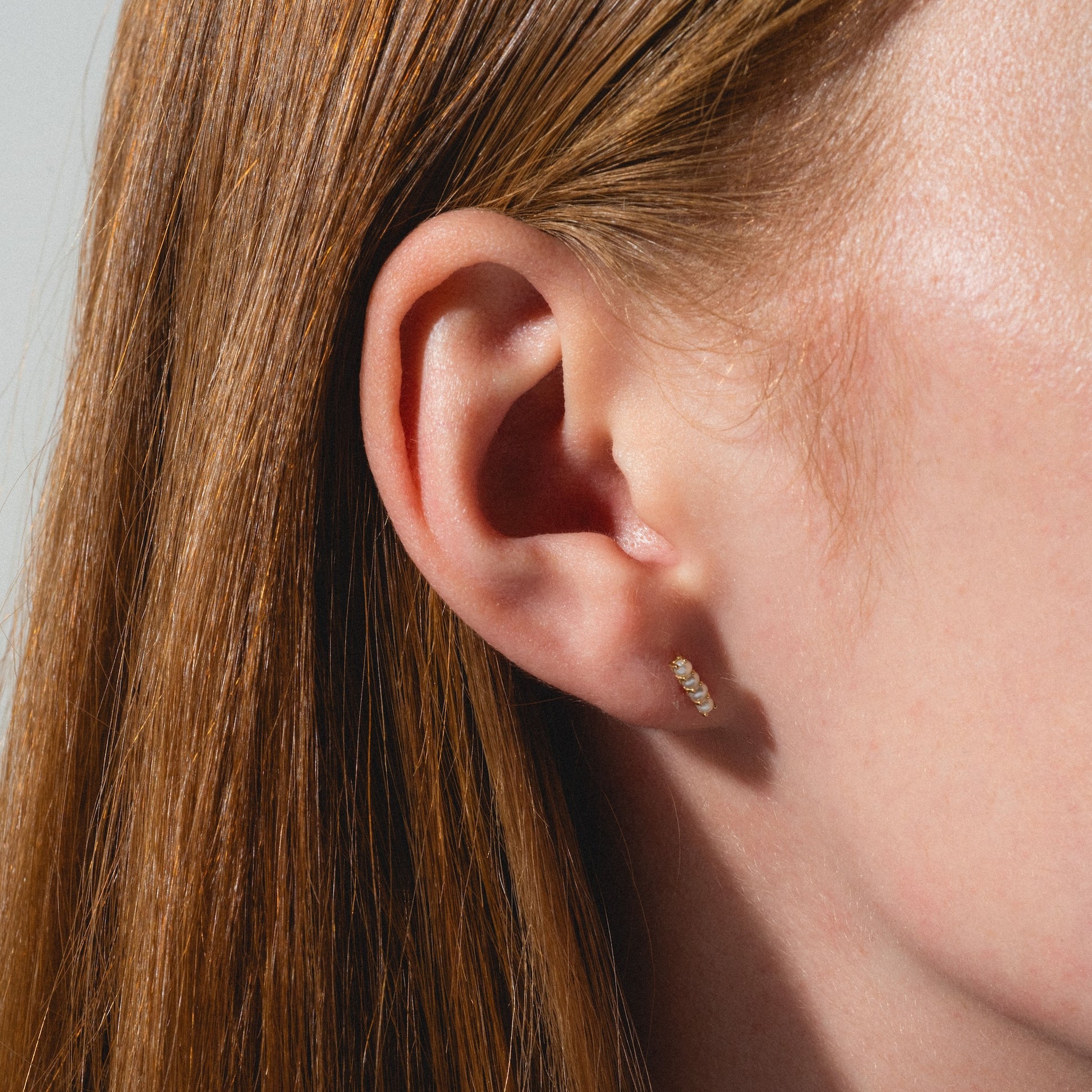 Close-up of a person's ear with straight, reddish-brown hair wearing Solid Gold Pearl Bar Flat Back Studs. The earring is small, gold, and bar-shaped with a pearl detail. The background is plain and neutral.