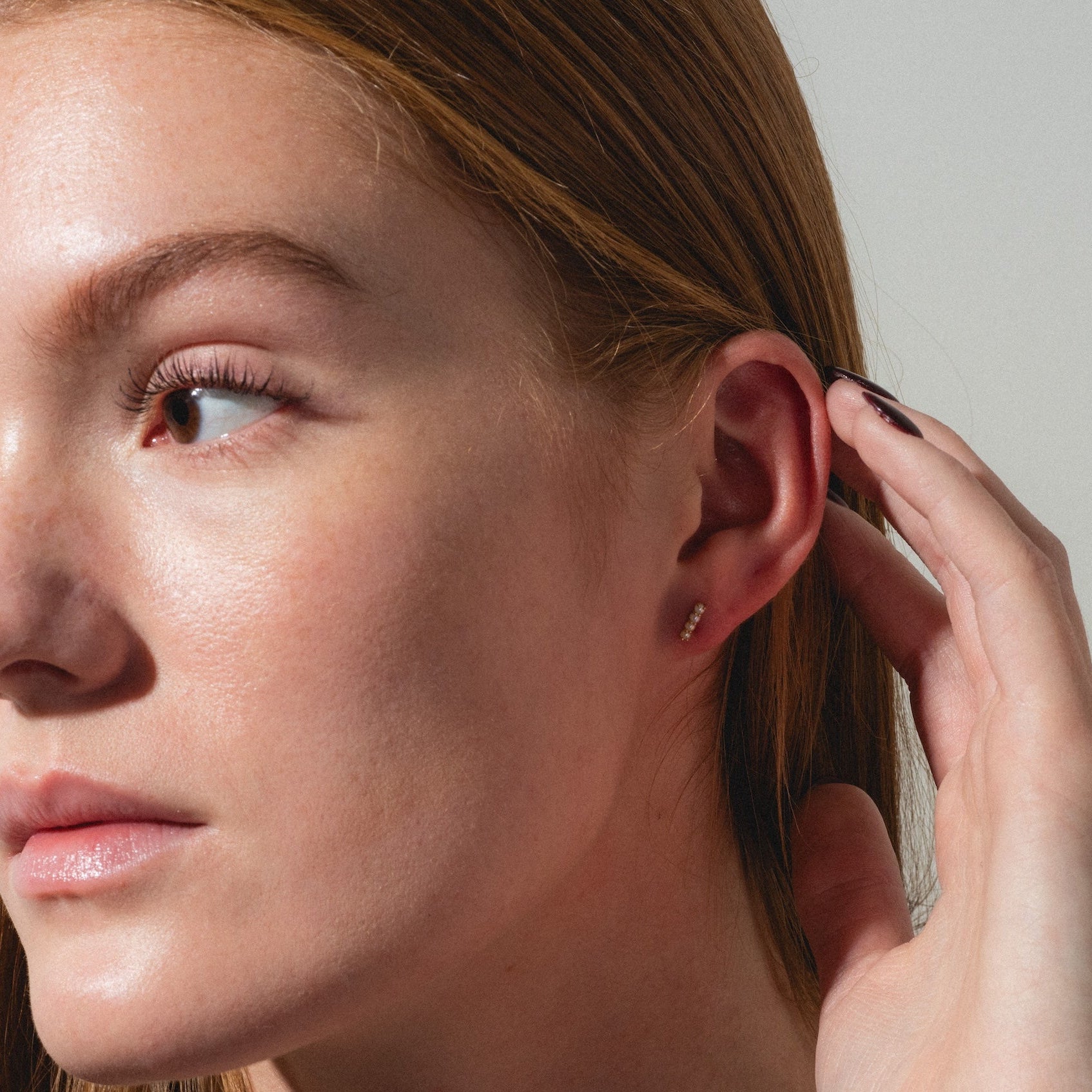 A close-up of a person with long red hair tucking their hair behind their ear, revealing Solid Gold Pearl Bar Flat Back Studs. The lighting highlights their clear skin and freckles.