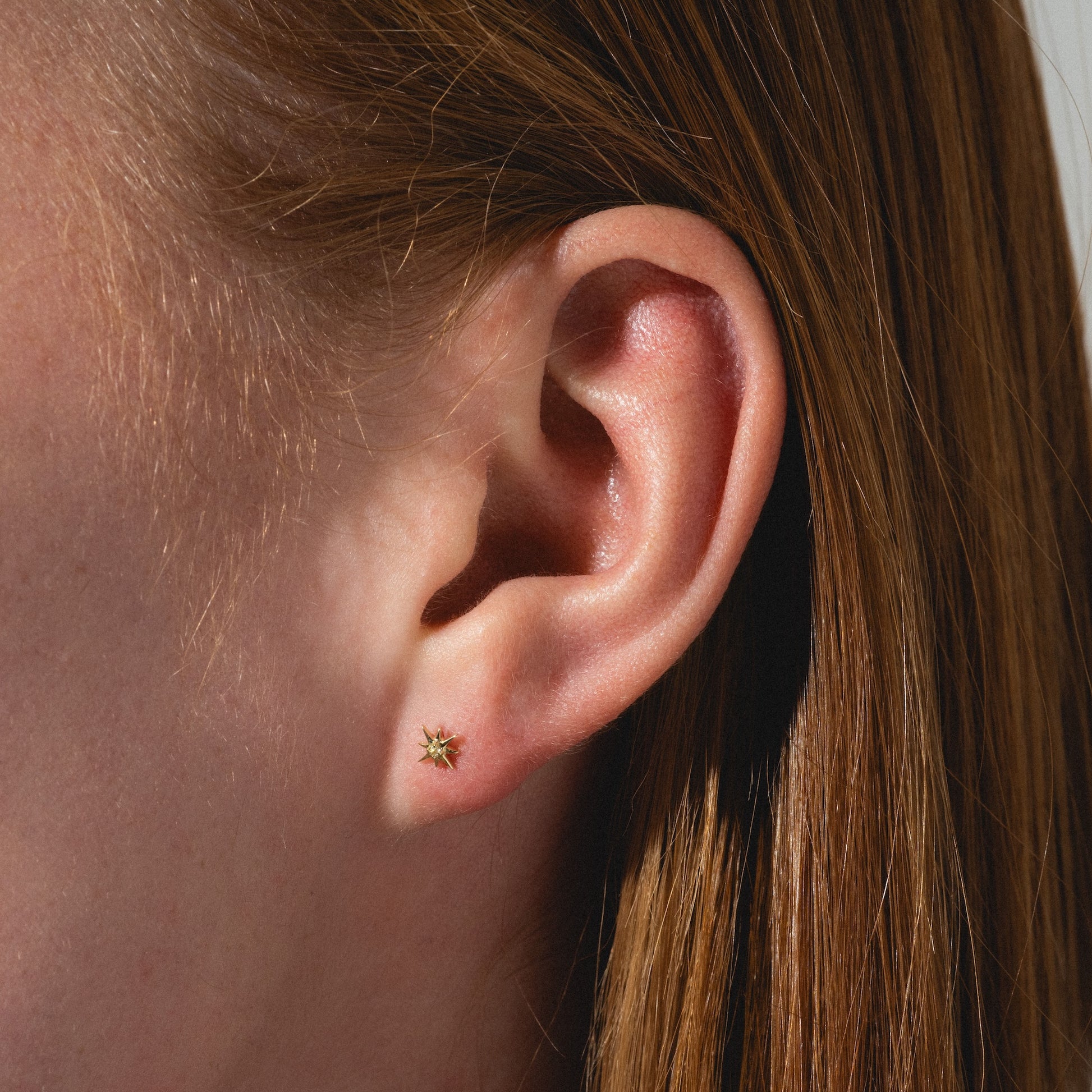 Close-up of a person’s ear with straight auburn hair, wearing the Solid Gold Starburst Flat Back Stud—a small gold star-shaped earring with a green gemstone center.