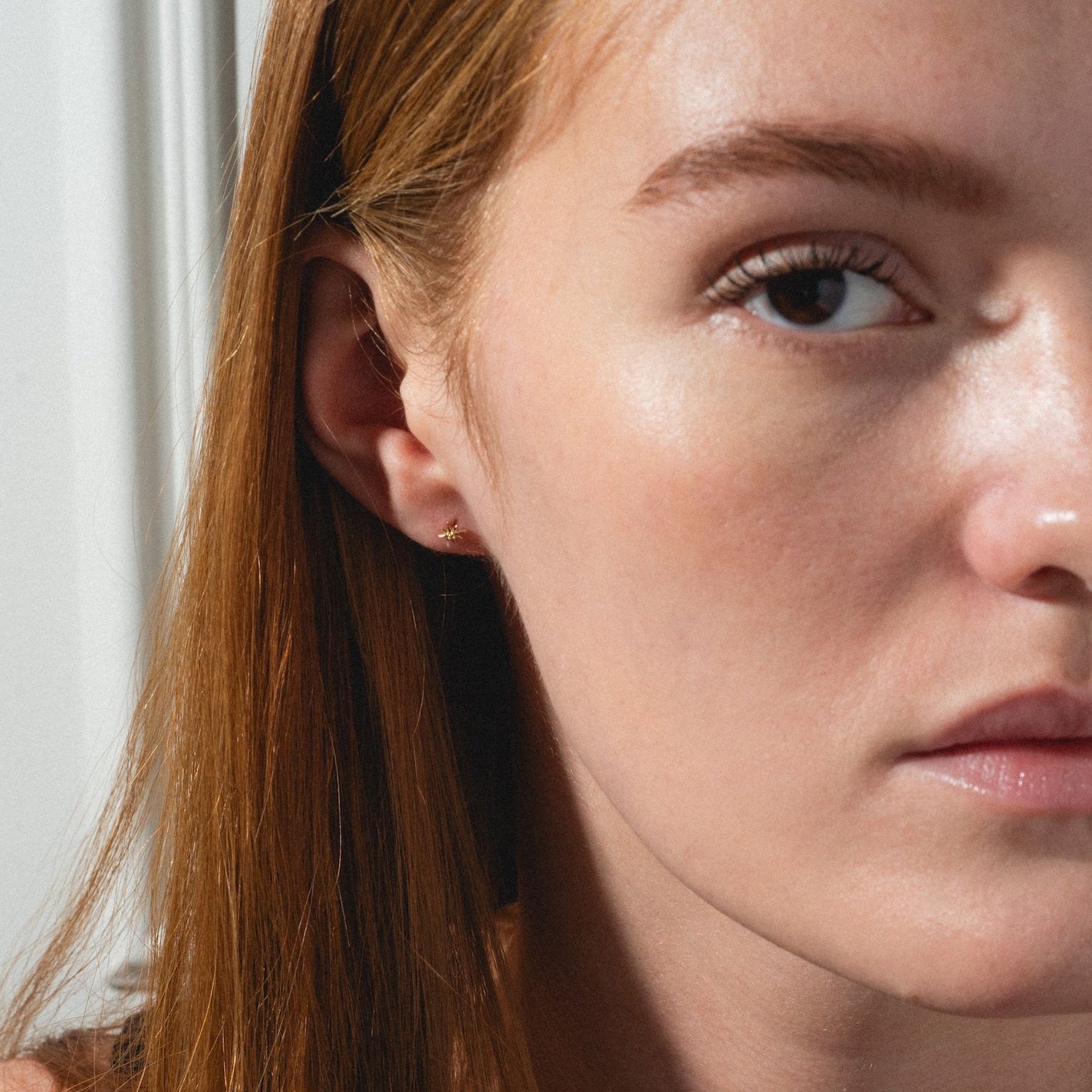A young woman with long red hair and fair skin stands in soft natural light near a white wall, wearing the Solid Gold Starburst Flat Back Studs and looking into the camera with a neutral expression.