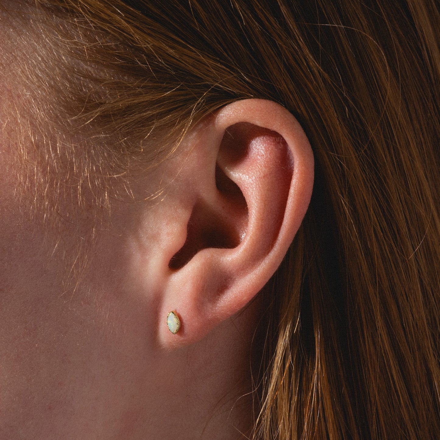 Close-up of a person's ear with straight auburn hair, wearing a Solid Gold Opal Marquise Flat Back Stud featuring a small marquise-shaped opal set in solid gold.