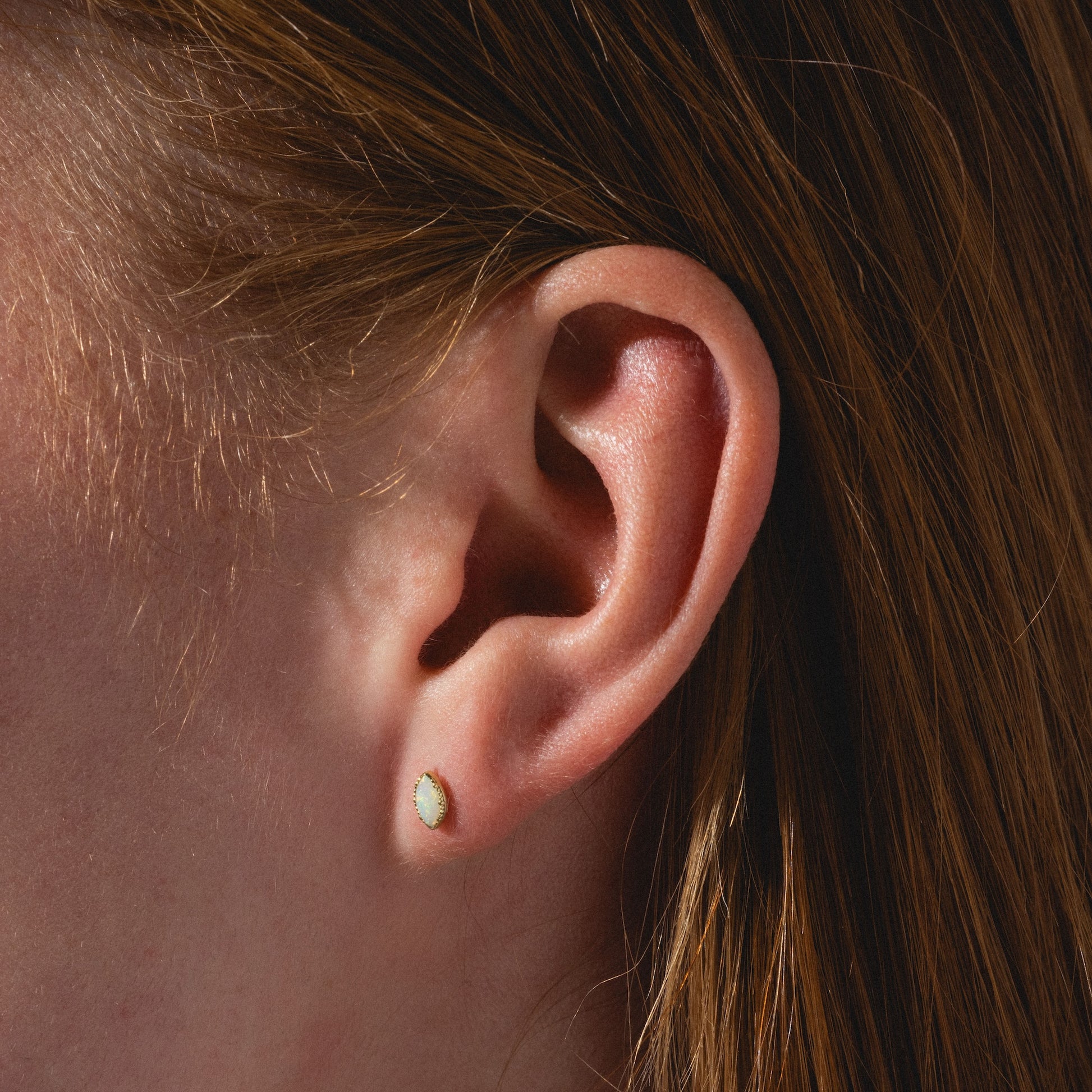 Close-up of a person's ear with straight auburn hair, wearing a Solid Gold Opal Marquise Flat Back Stud featuring a small marquise-shaped opal set in solid gold.