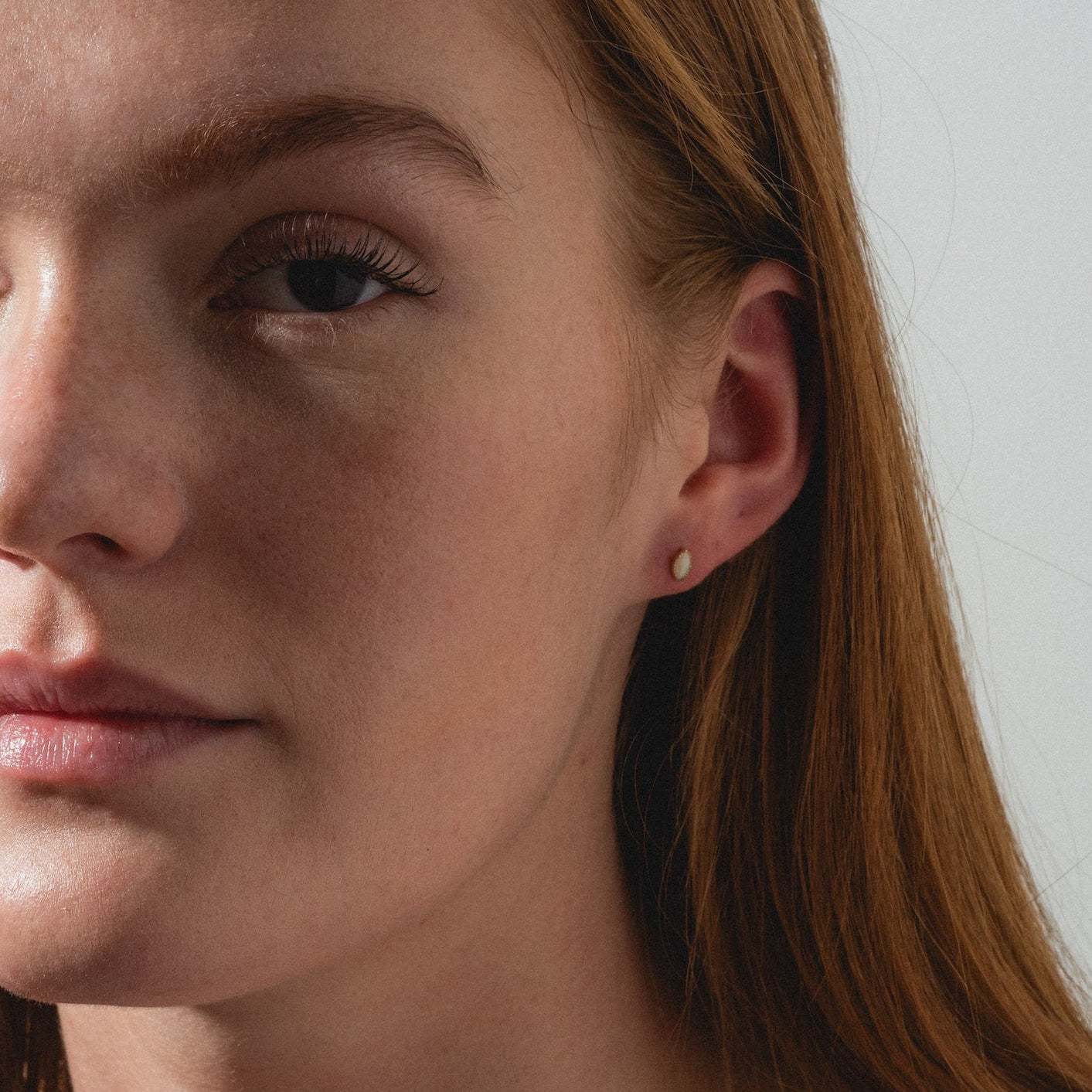 A woman with long red hair and fair skin, dressed in a black top, gazes slightly past the camera indoors, showcasing Solid Gold Opal Marquise Flat Back Studs.