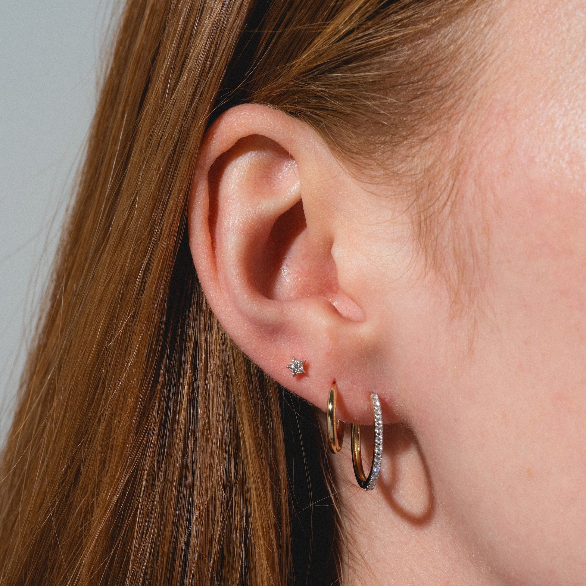 Close-up of a person's ear with three piercings: a Solid Gold Sparkling Star Flat Back Stud in the upper lobe, and two gold hoops—one plain, one with small stones—in the lower lobe. The person has straight, light brown hair.