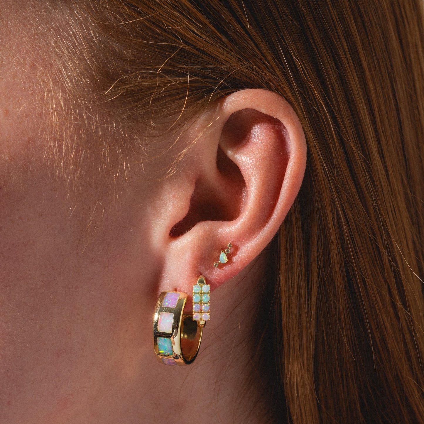 Close-up of a person’s ear with three piercings, each featuring Solid Gold Opal Constellation Flat Back Studs. The gold earrings have iridescent opal stones; the person has light skin and straight reddish-brown hair.