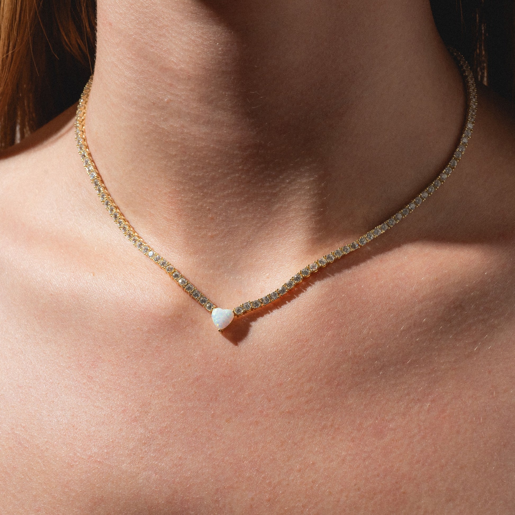A close-up of a person's neck wearing the Queen of Hearts Tennis Necklace, featuring a delicate gold chain with a small, heart-shaped white stone pendant at the center.