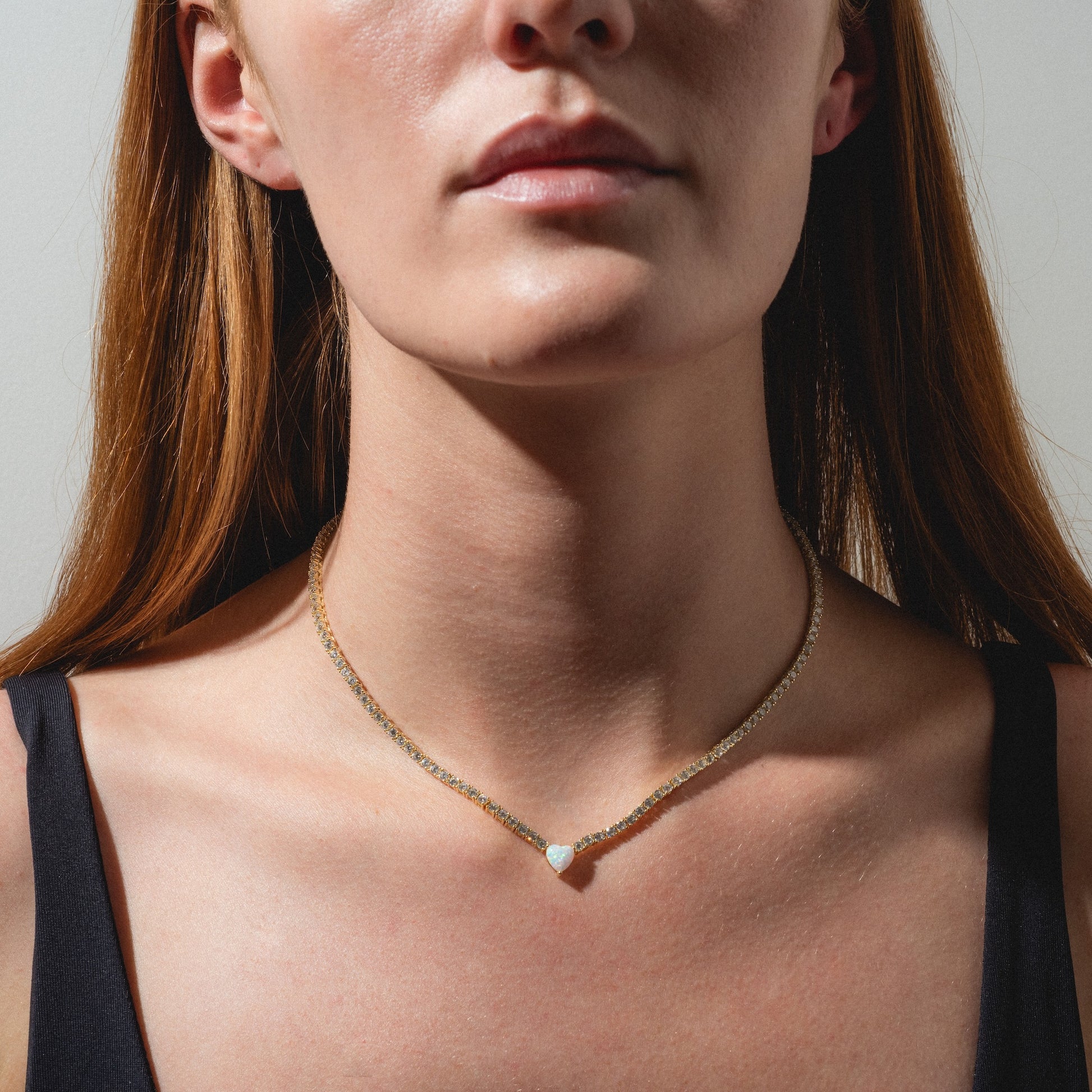A woman with long brown hair wears the Queen of Hearts Tennis Necklace—a delicate gold chain with a teardrop-shaped pendant. She is dressed in a black top and shown from the shoulders up against a plain background.