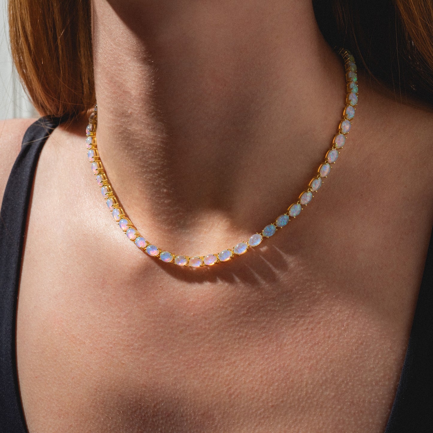 A close-up of a woman’s neck wearing the Baby Blue Opal Eternal Tennis Necklace, featuring round opal stones, against her skin and a black top with sunlight highlighting the necklace.