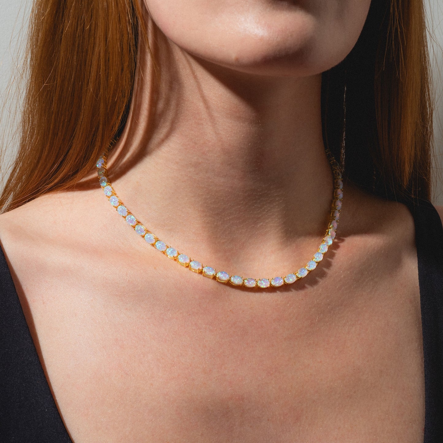 A woman wearing a black top models the Baby Blue Opal Eternal Tennis Necklace, featuring round, iridescent blue opals that rest elegantly on her neck and collarbone.