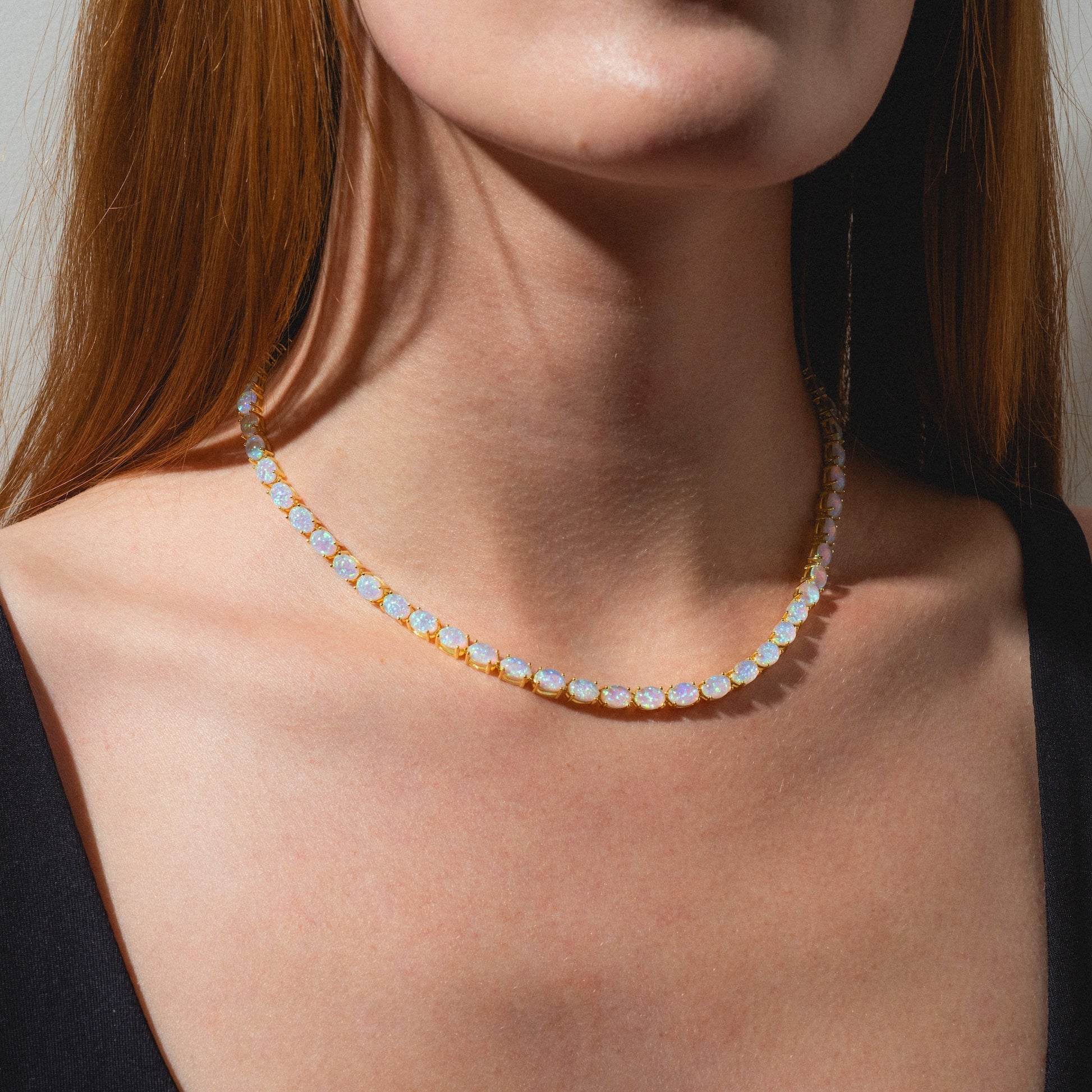 A woman wearing a black top models the Baby Blue Opal Eternal Tennis Necklace, featuring round, iridescent blue opals that rest elegantly on her neck and collarbone.