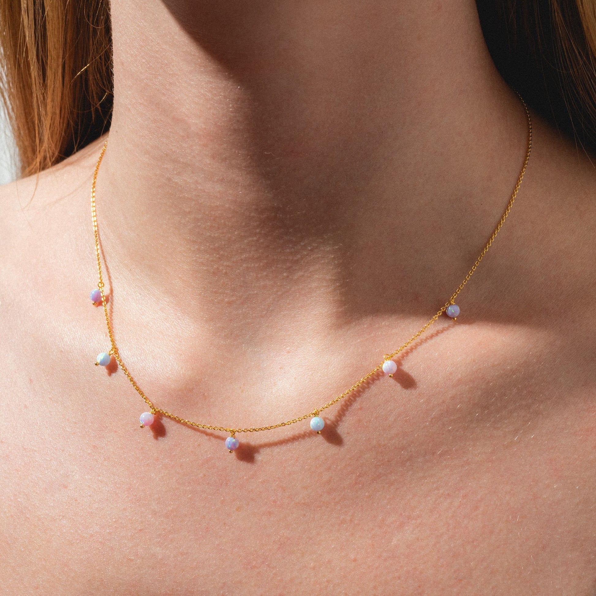 A close-up of a person’s neck wearing the Cotton Candy Opal Droplet Necklace— a delicate gold chain adorned with evenly spaced, small pastel beads. The softly lit background is minimal.
