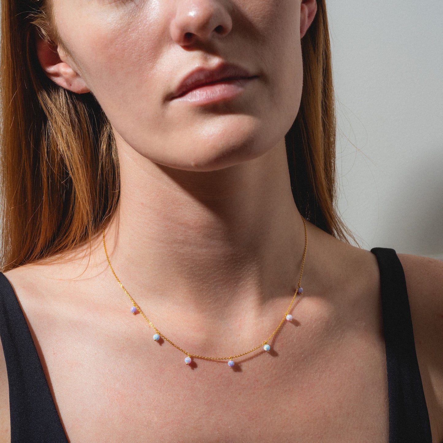 A woman wears a black top and the Cotton Candy Opal Droplet Necklace—a delicate gold piece with small, multicolored beads—photographed from the shoulders up against a plain background.