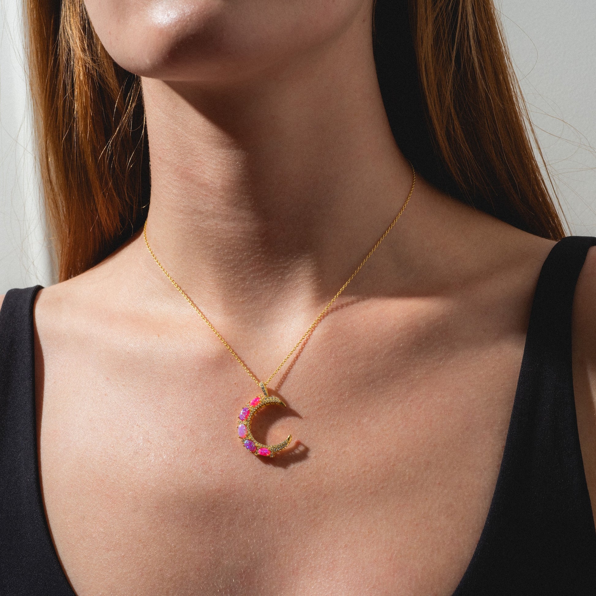 A woman wearing a black top and the Pink Opal Crescent Moon Pendant, shown from shoulders to chin in bright natural light.