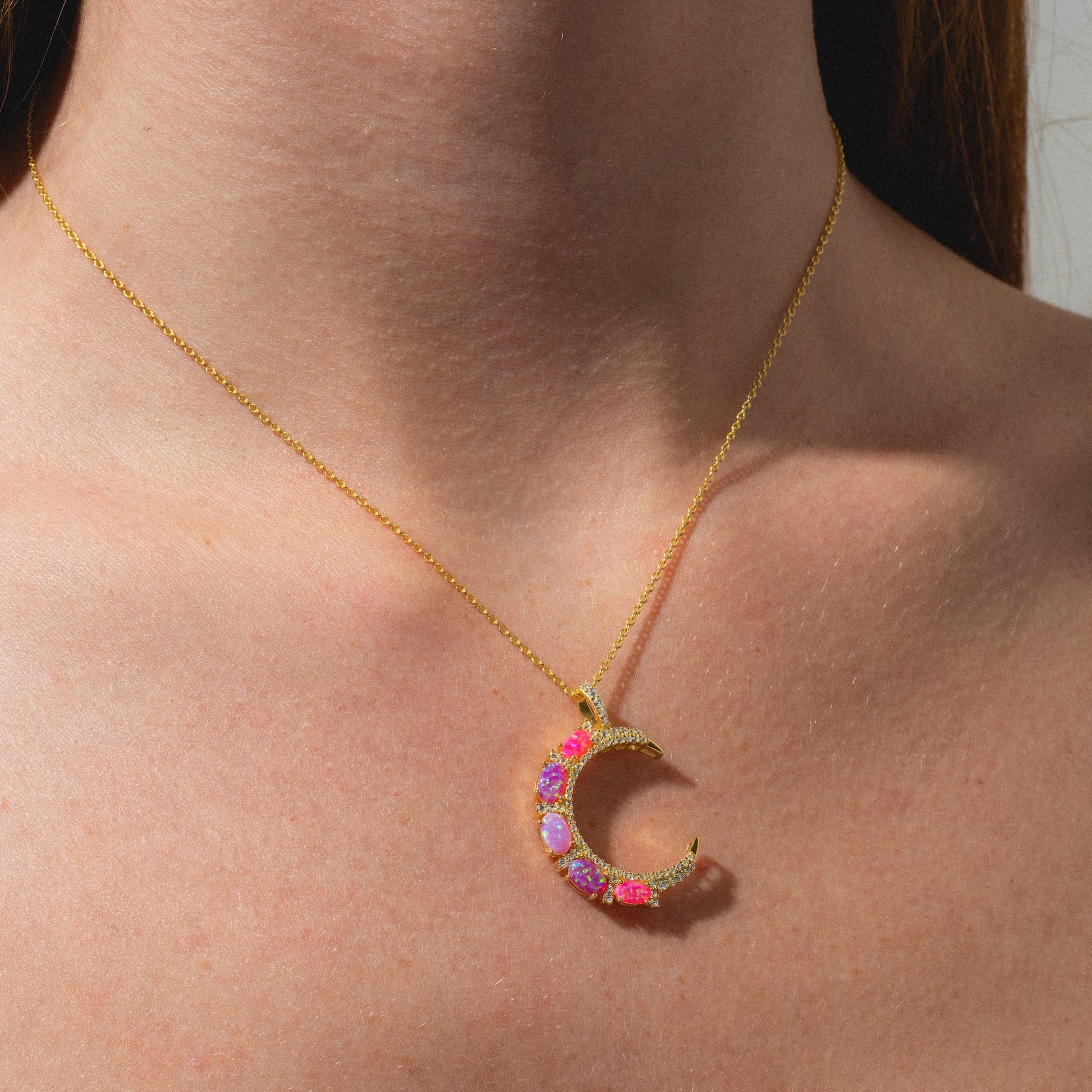 A person wearing the Pink Opal Crescent Moon Pendant, a gold chain necklace featuring a crescent moon adorned with pink and purple gemstones, shown on bare skin with part of their brown hair visible.