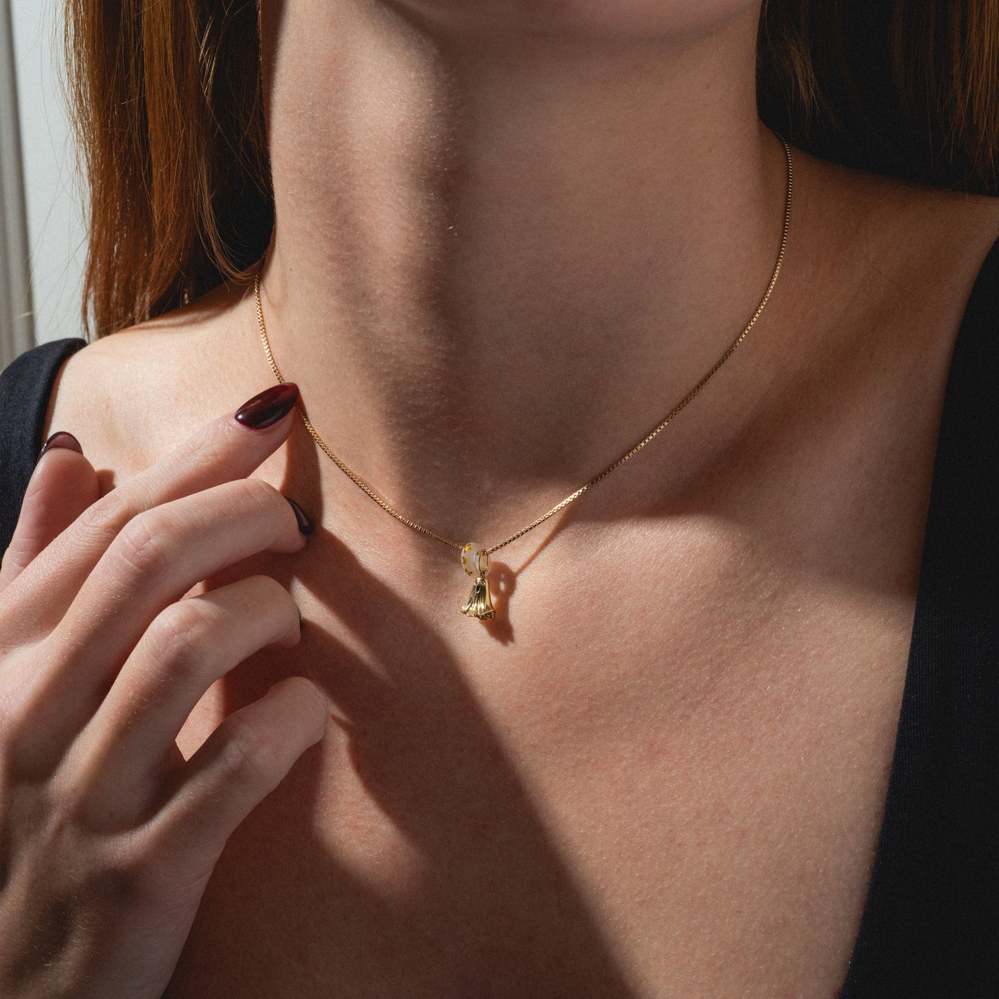 A woman in a black top gently touches the Limited Edition Solid Gold Halloween Superstitions Charm on her neck, her dark red nails visible as the image highlights the elegant necklace against her collarbone.