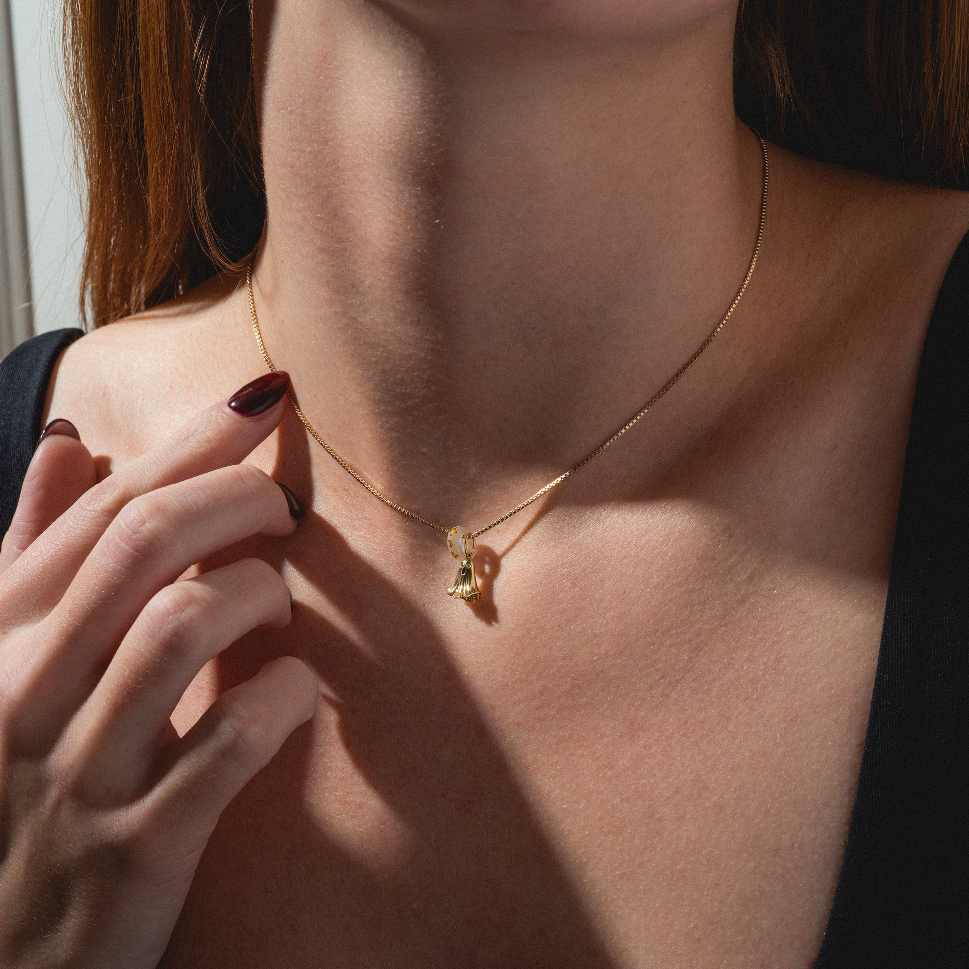 A woman in a black top gently touches the Limited Edition Solid Gold Halloween Superstitions Charm on her neck, her dark red nails visible as the image highlights the elegant necklace against her collarbone.