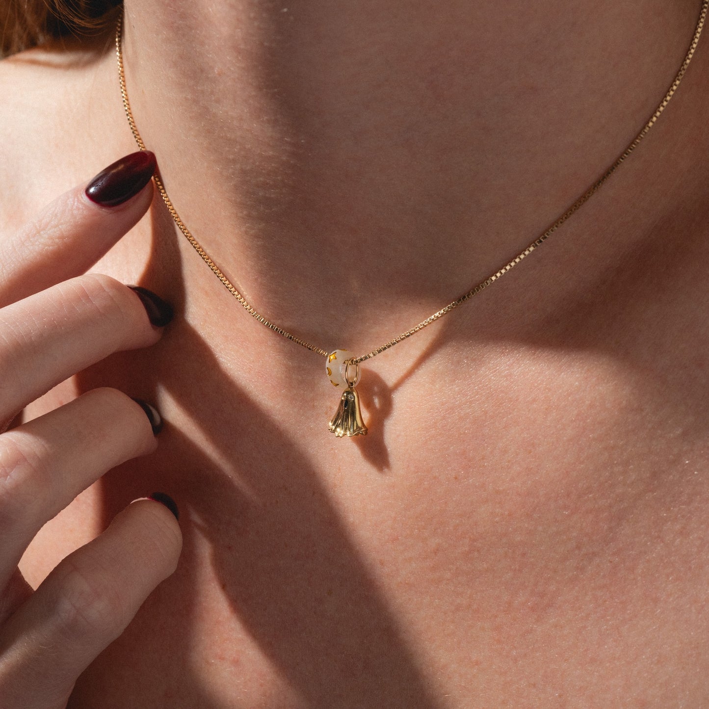 A close-up of a person’s neck wearing the Limited Edition Solid Gold Halloween Superstitions Charm, featuring two delicate charms—a bell and a pearl. The person's hand with dark red nail polish touches the necklace.