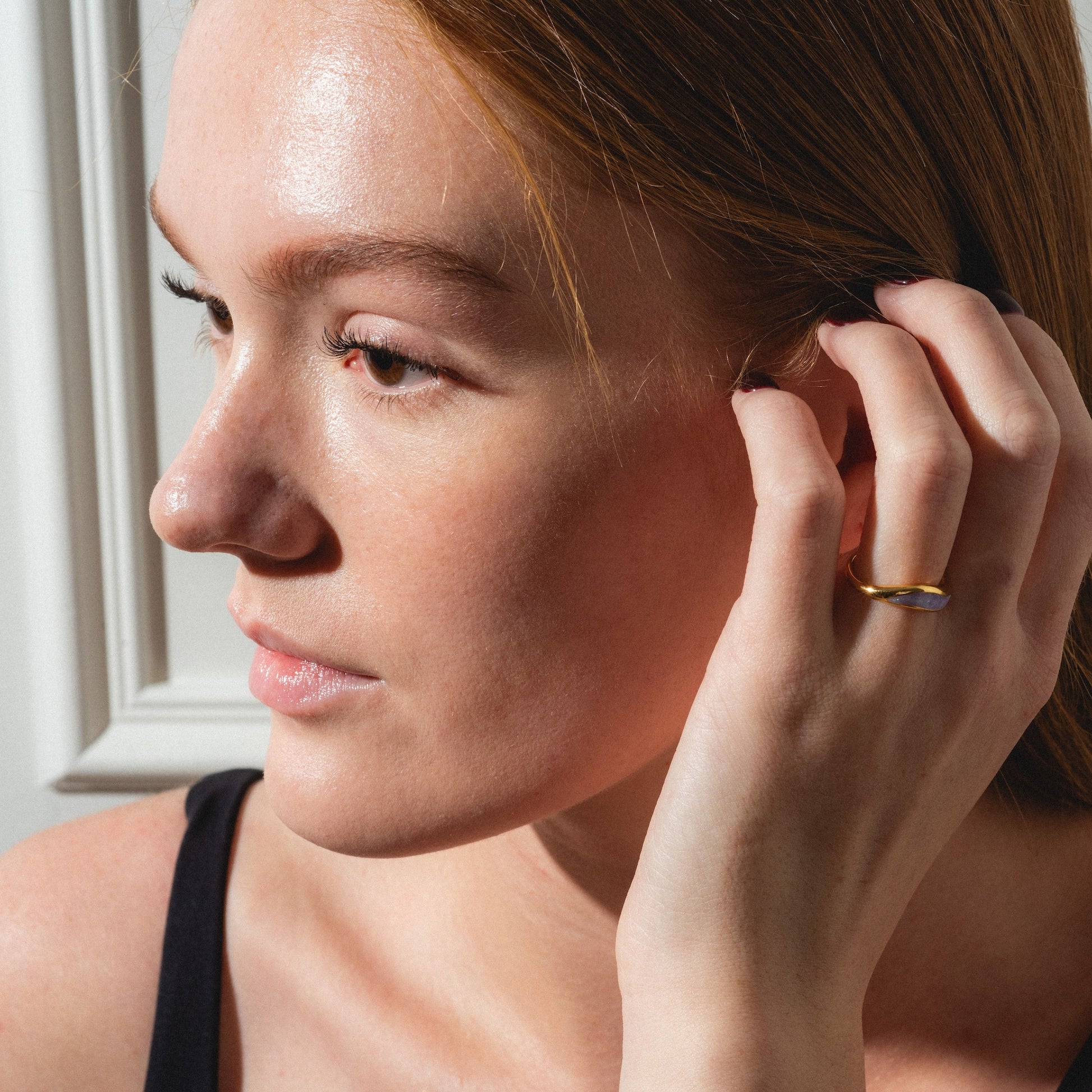 A woman with fair skin and auburn hair touches her ear, wearing the Opal Dreamers Wave Ring—a gold ring set with a purple stone. She wears a black top, illuminated by natural sunlight.
