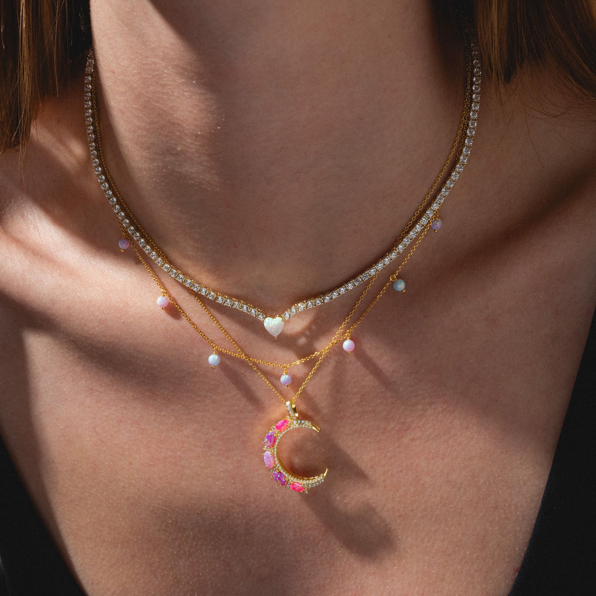 A close-up of a woman's neck wearing three layered necklaces: a diamond choker, a gold chain with iridescent beads, and the Cotton Candy Opal Droplet Necklace featuring a gold crescent moon pendant adorned with pink and white stones.