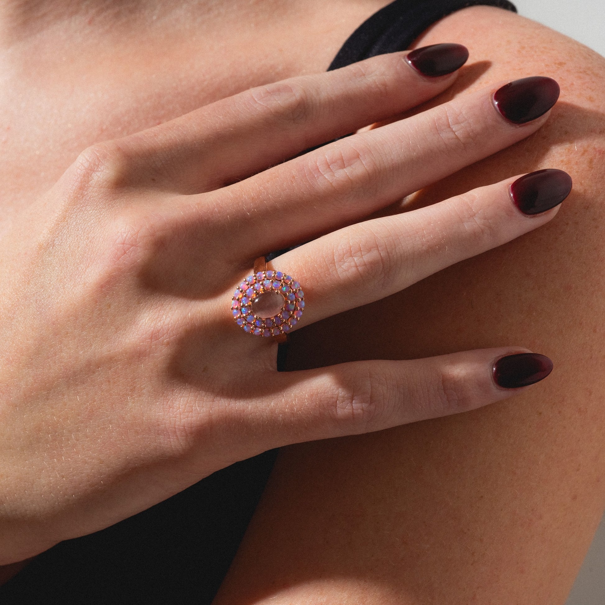 A hand with dark burgundy nails rests on a shoulder, wearing the Rose Gold Pink Opal and Cats Eye Moonstone Crown Ring.