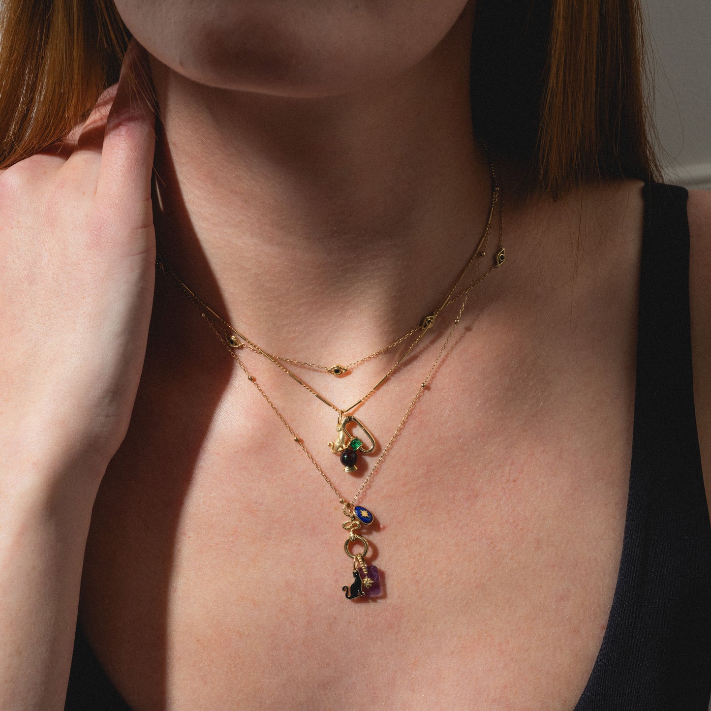 A woman wears the Limited Edition Solid Gold Halloween Superstitions Charm layered on gold necklaces. Dressed in a black top, she softly touches her neck as gentle lighting highlights the unique charm and casts subtle shadows.