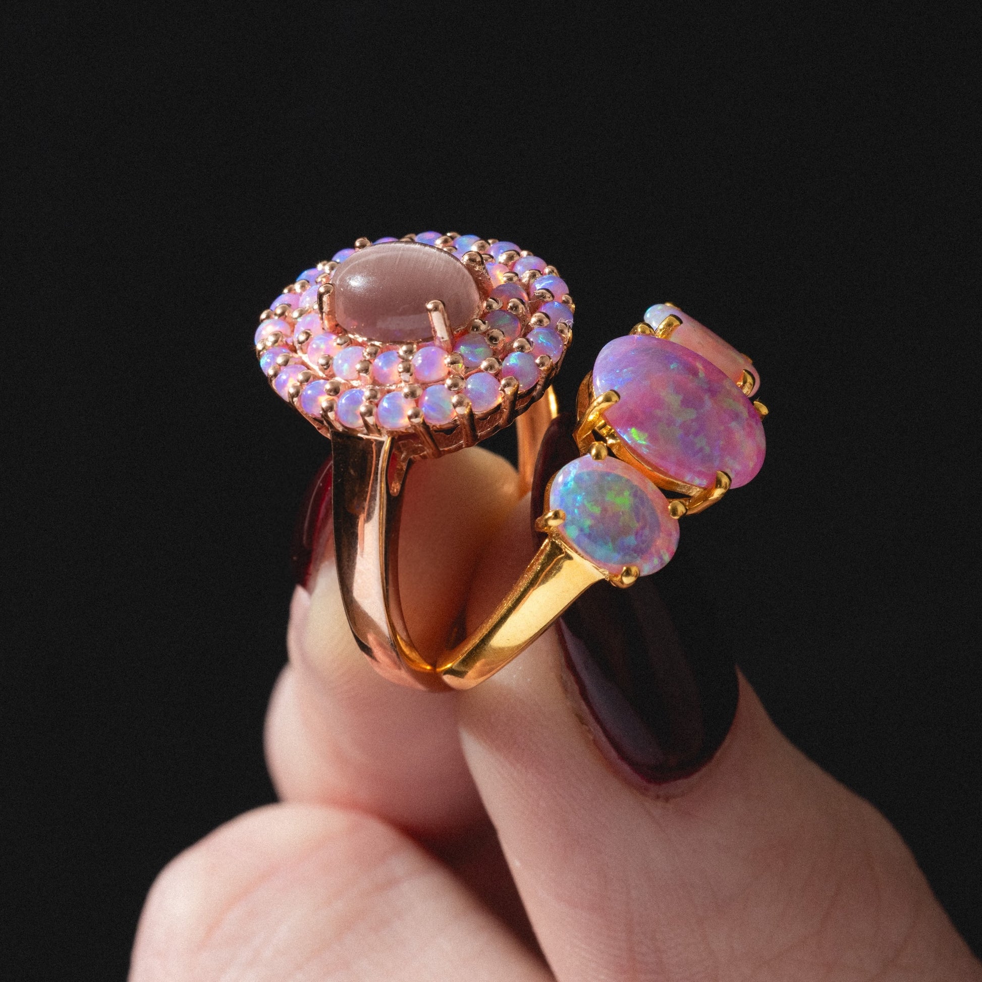 A hand with dark red nail polish holds two Pink Opal Gum Drop Rings, each set with pink and opalescent gemstones, against a black background.