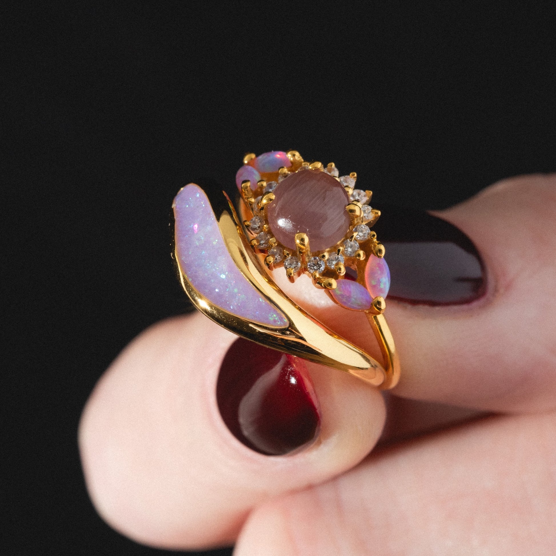 A hand with dark red nail polish holds the Opal Dreamers Wave Ring—a gold band with a large oval gemstone encircled by smaller stones and an iridescent opal accent—against a black background.