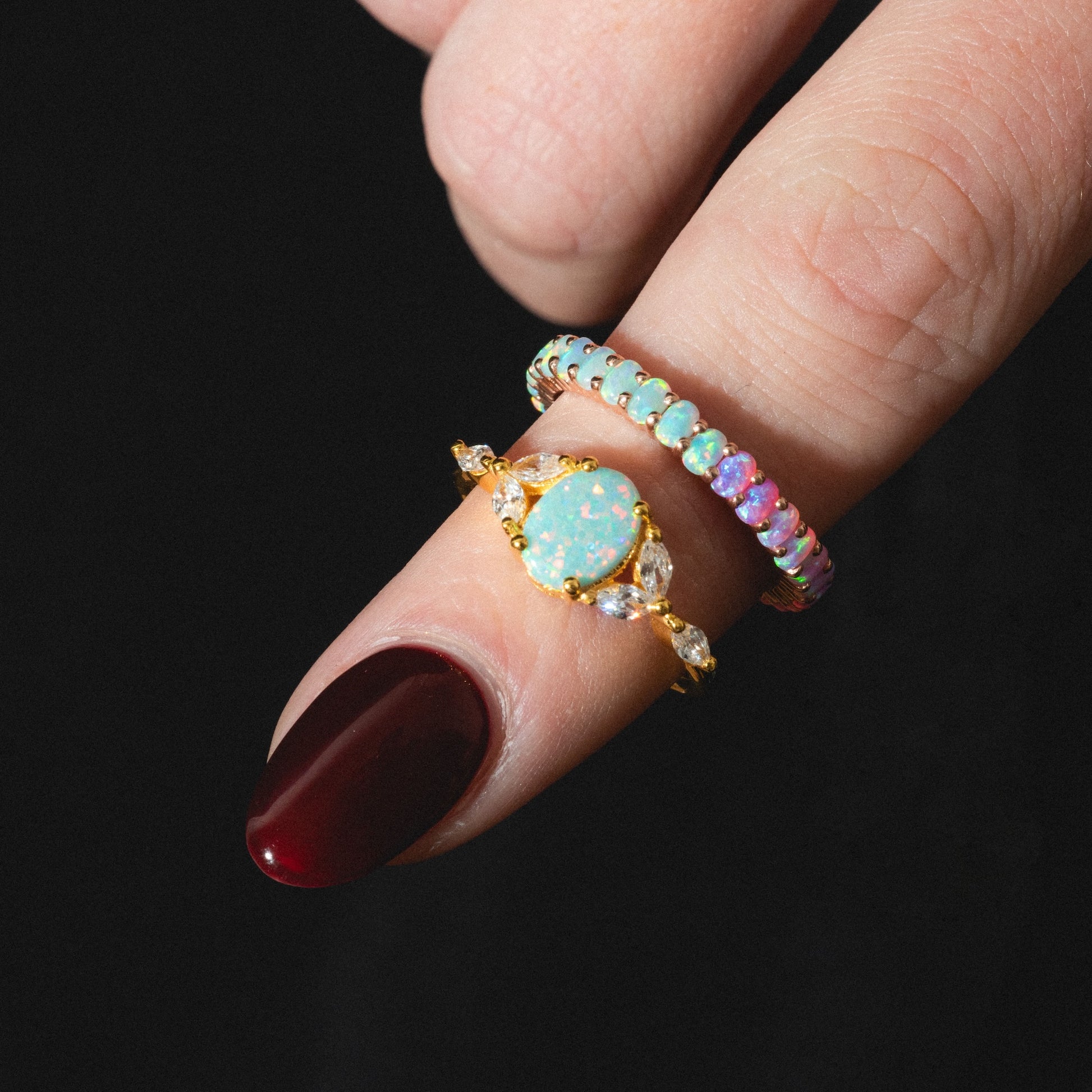 A hand with dark red nail polish wears two rings: the Opal Enchanted Ring—gold with an oval opal and clear stones—and a band of small multicolored opals, set against a black background.