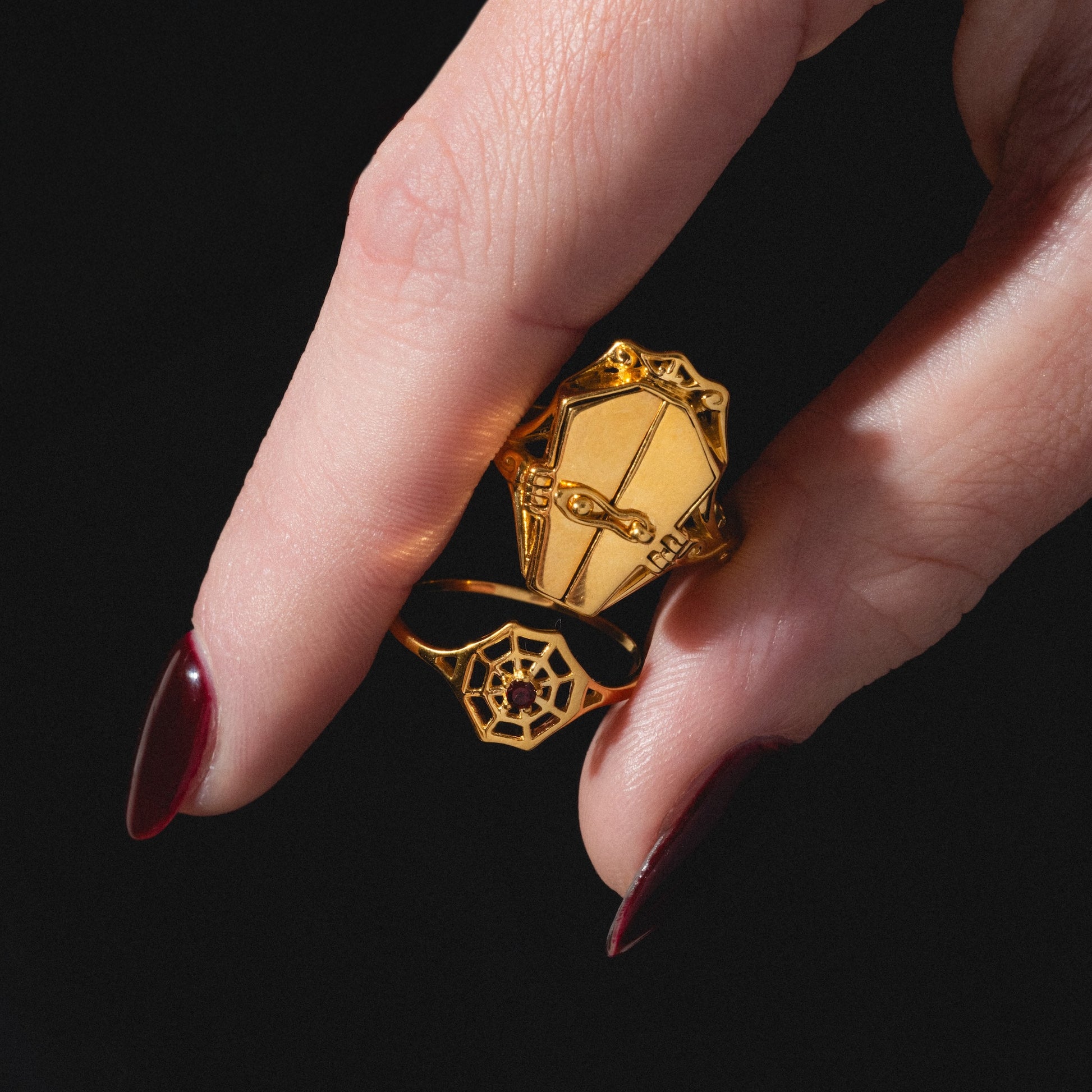 A hand with dark red nails holds two gold rings: one shaped like a coffin with a cross, and the other is the Limited Edition Black Widow Garnet Spiderweb Ring, featuring a spiderweb design with a small red gem in the center. The background is black.