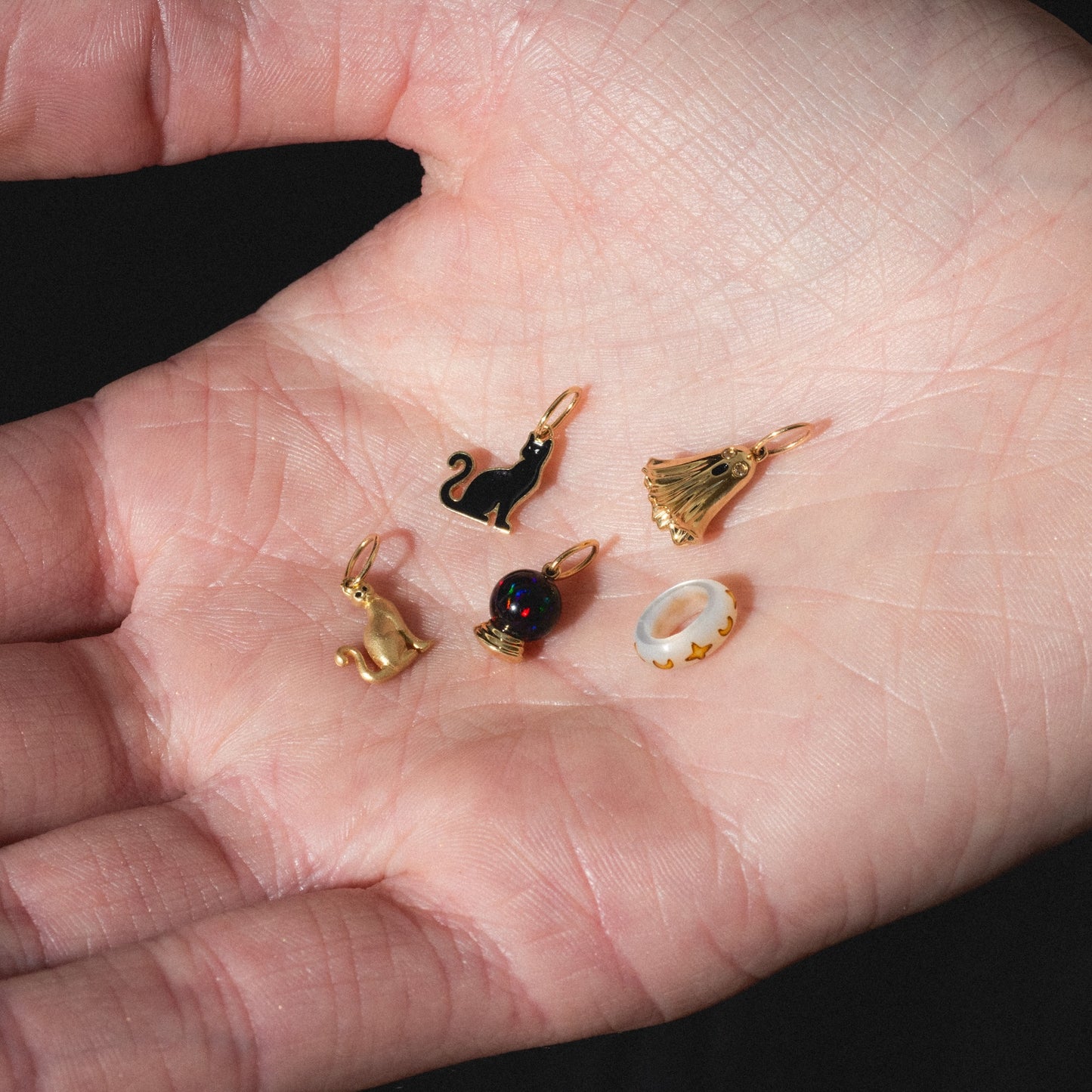 A hand holds five Limited Edition Solid Gold Halloween Superstitions Charms: a black cat, gold cat, black round bead, gold ghost, and a white oval charm with gold crescent moons.