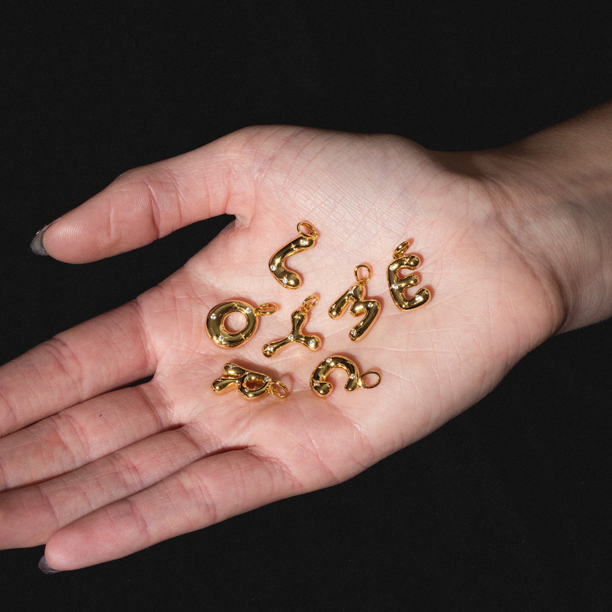 A hand with palm facing up holds several shiny gold-colored Star Crossed Celestial Bubble Letter Charms shaped like abstract squiggles and curves against a black background.