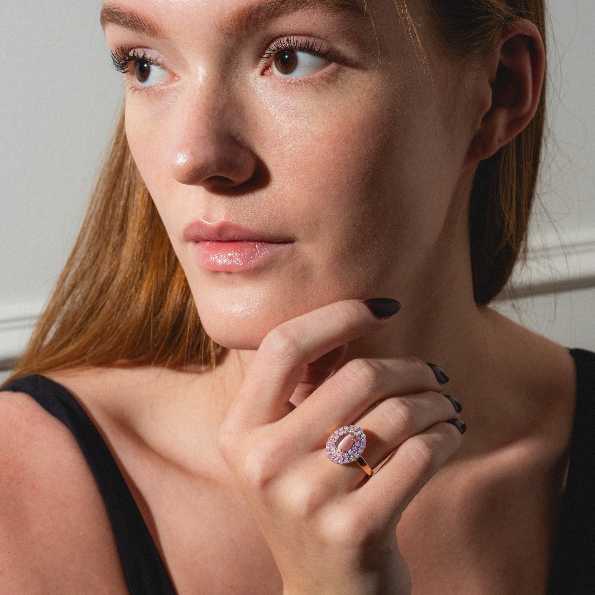 A woman with long auburn hair in a sleeveless black top gazes aside, showcasing the Rose Gold Pink Opal and Cats Eye Moonstone Crown Ring on her finger. Light accentuates her clear complexion and the ring's pink and white stones.