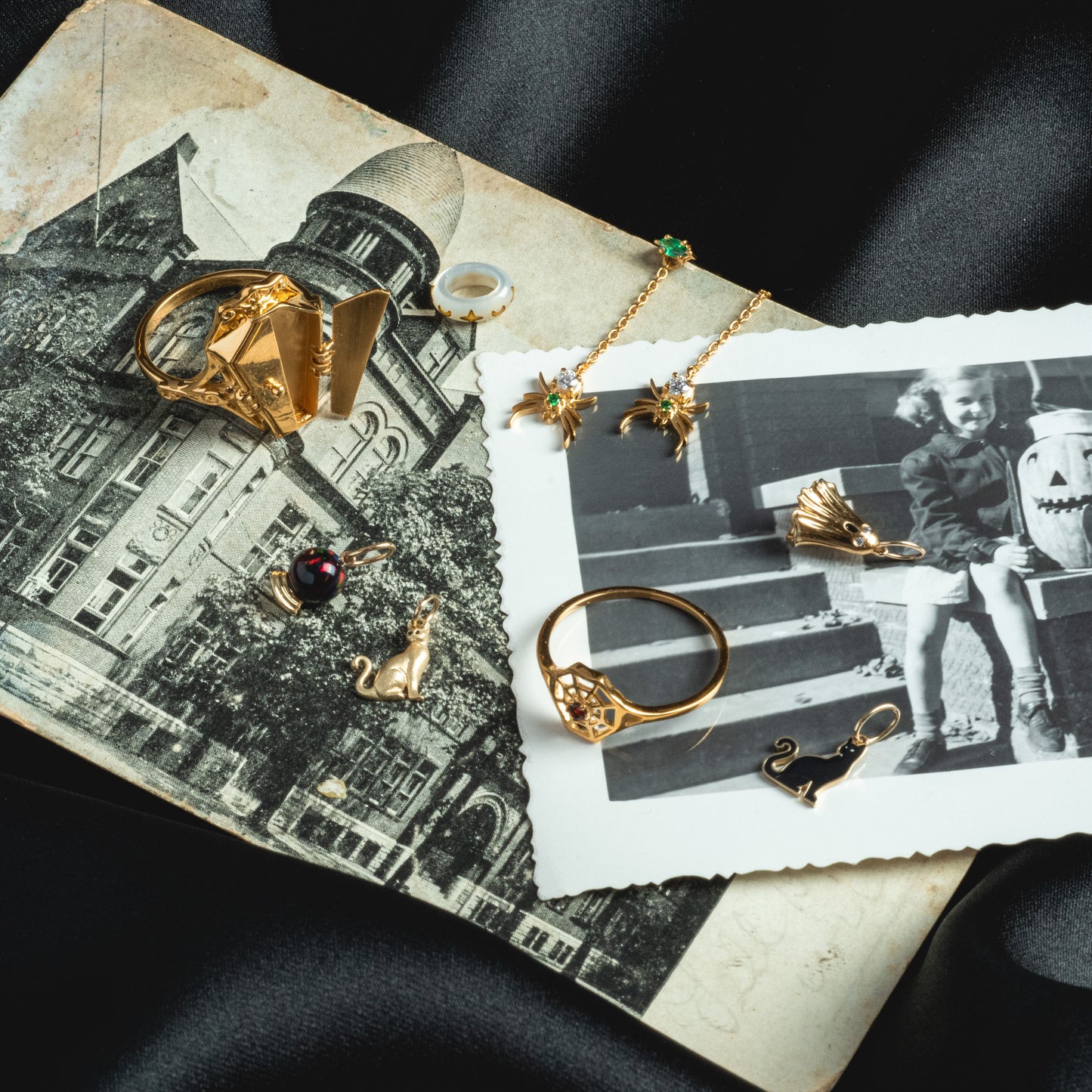 The Limited Edition Solid Gold Halloween Superstitions Charm is displayed with gold rings and a necklace on black-and-white photos of a historic building and a child with a carved pumpkin, all atop silky black fabric.