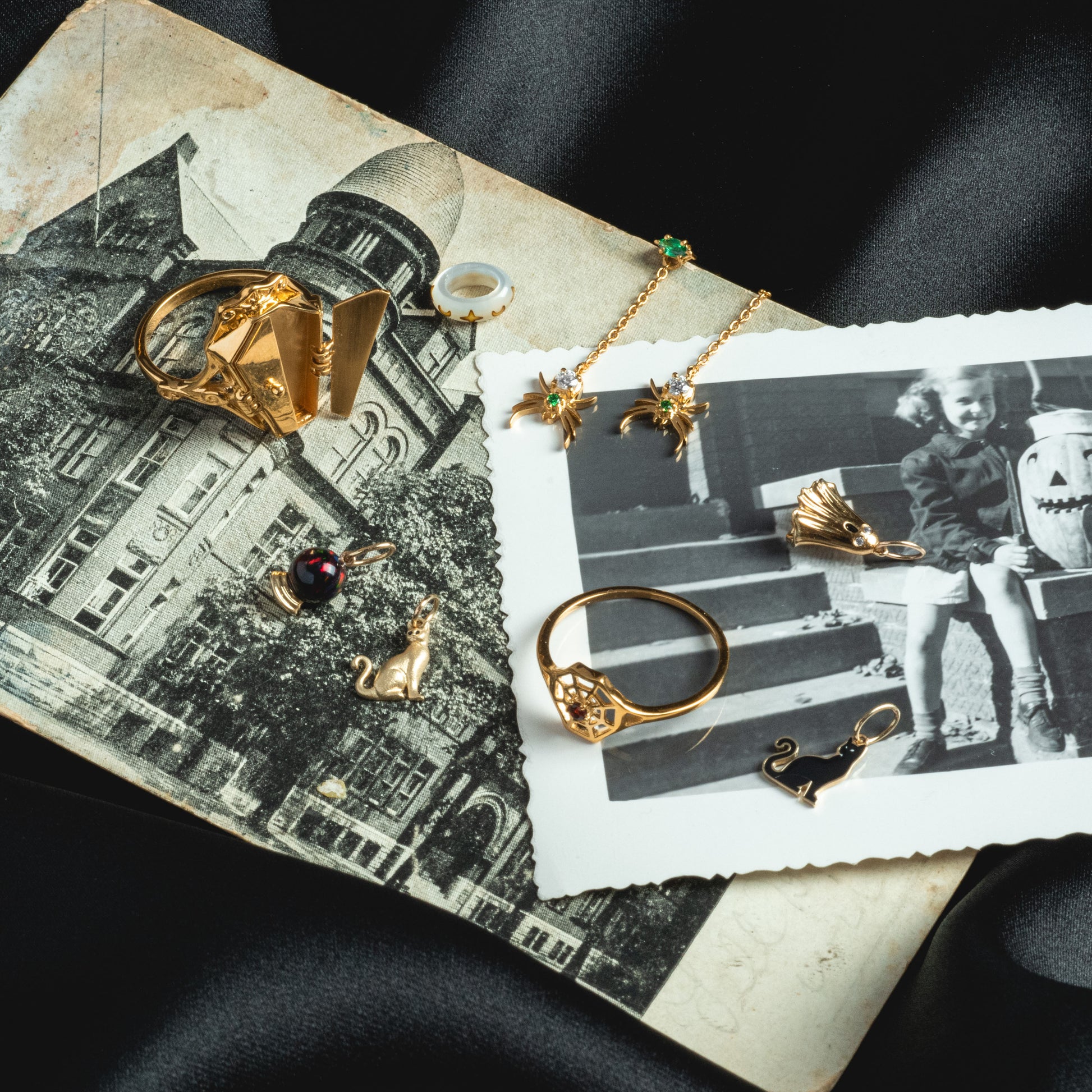 The Limited Edition Solid Gold Halloween Superstitions Charm is displayed with gold rings and a necklace on black-and-white photos of a historic building and a child with a carved pumpkin, all atop silky black fabric.